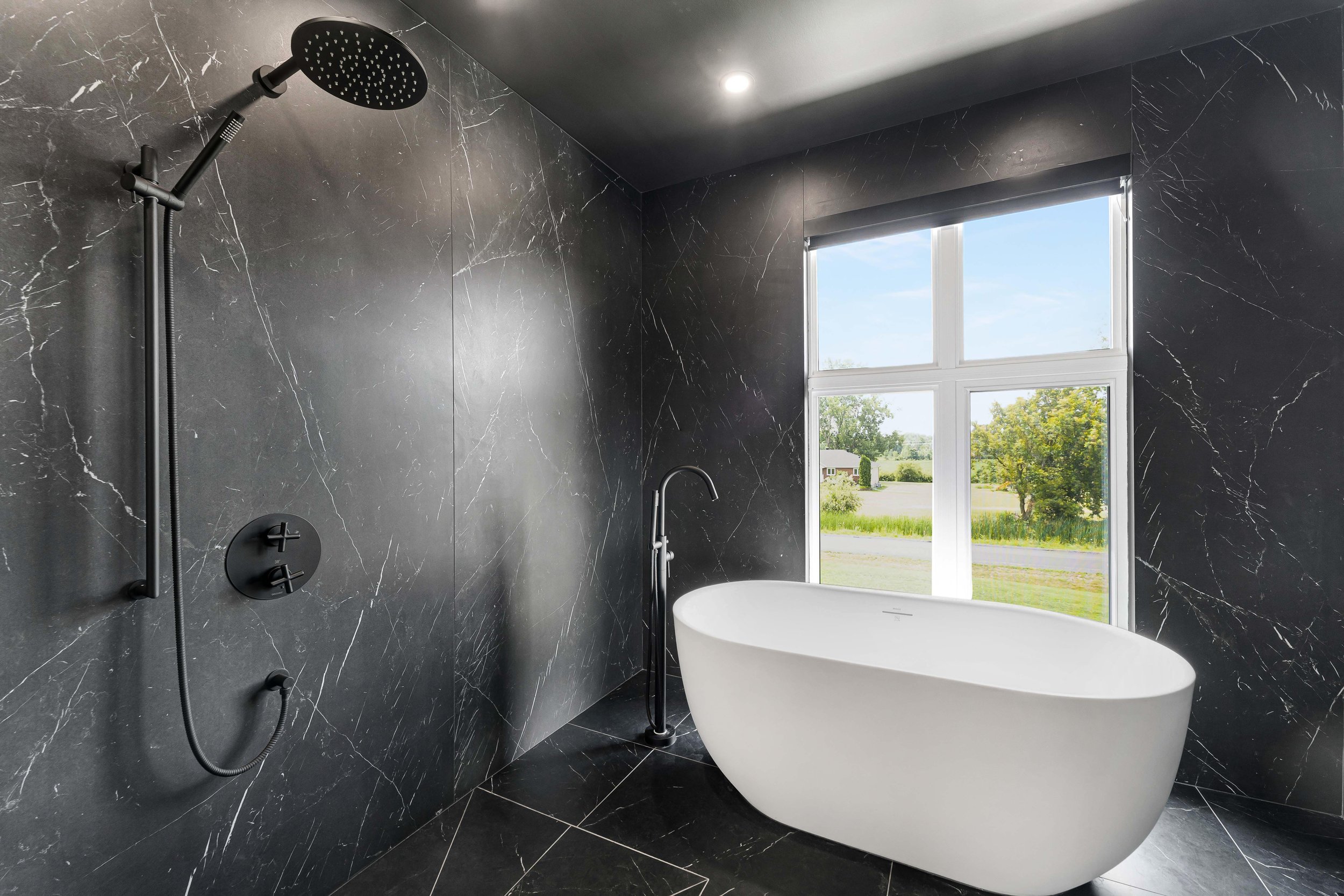 Modern bathroom with black marble walls, large freestanding white bathtub, black fixtures, and a window with a countryside view.