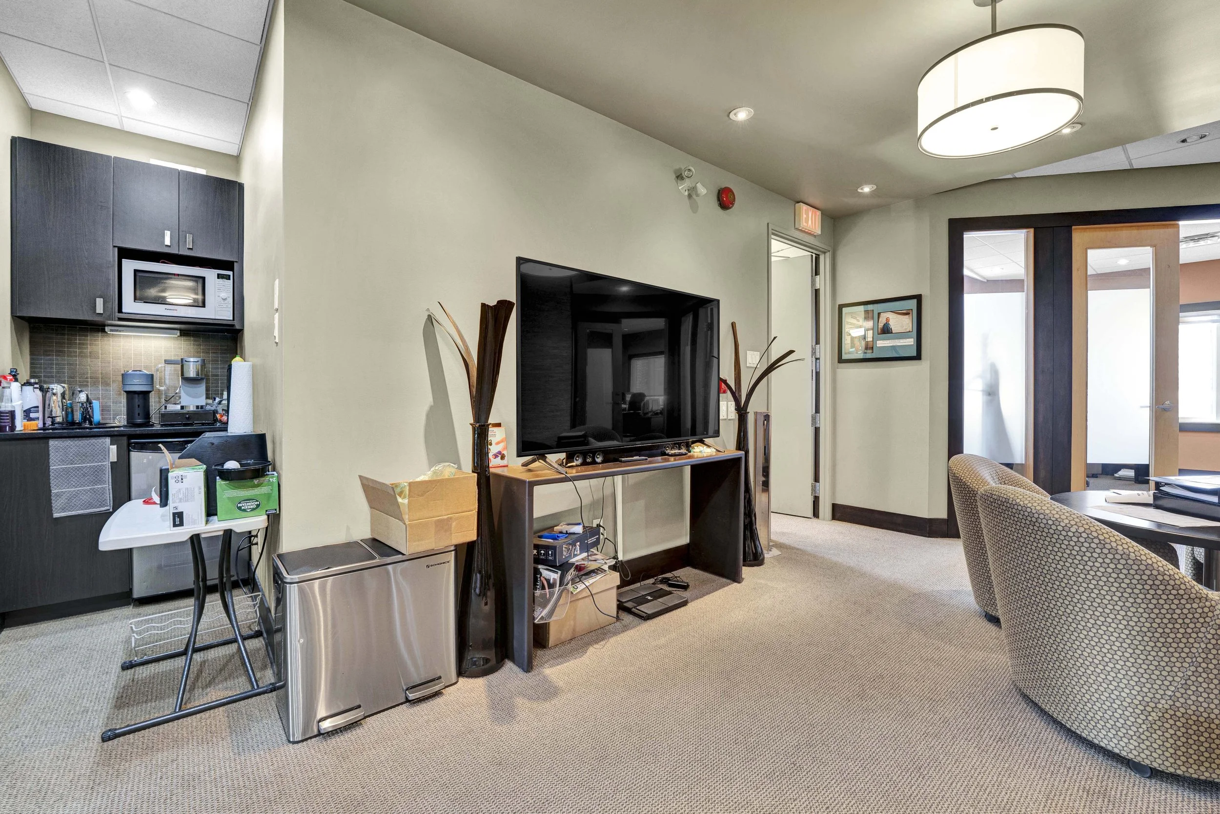 OttInside an office with a television, a kitchenette area, and a seating area. Items on the counter include paper towels, a coffee maker, and various bottles. The room has beige walls, a carpeted floor, and a ceiling with a large round light fixture.