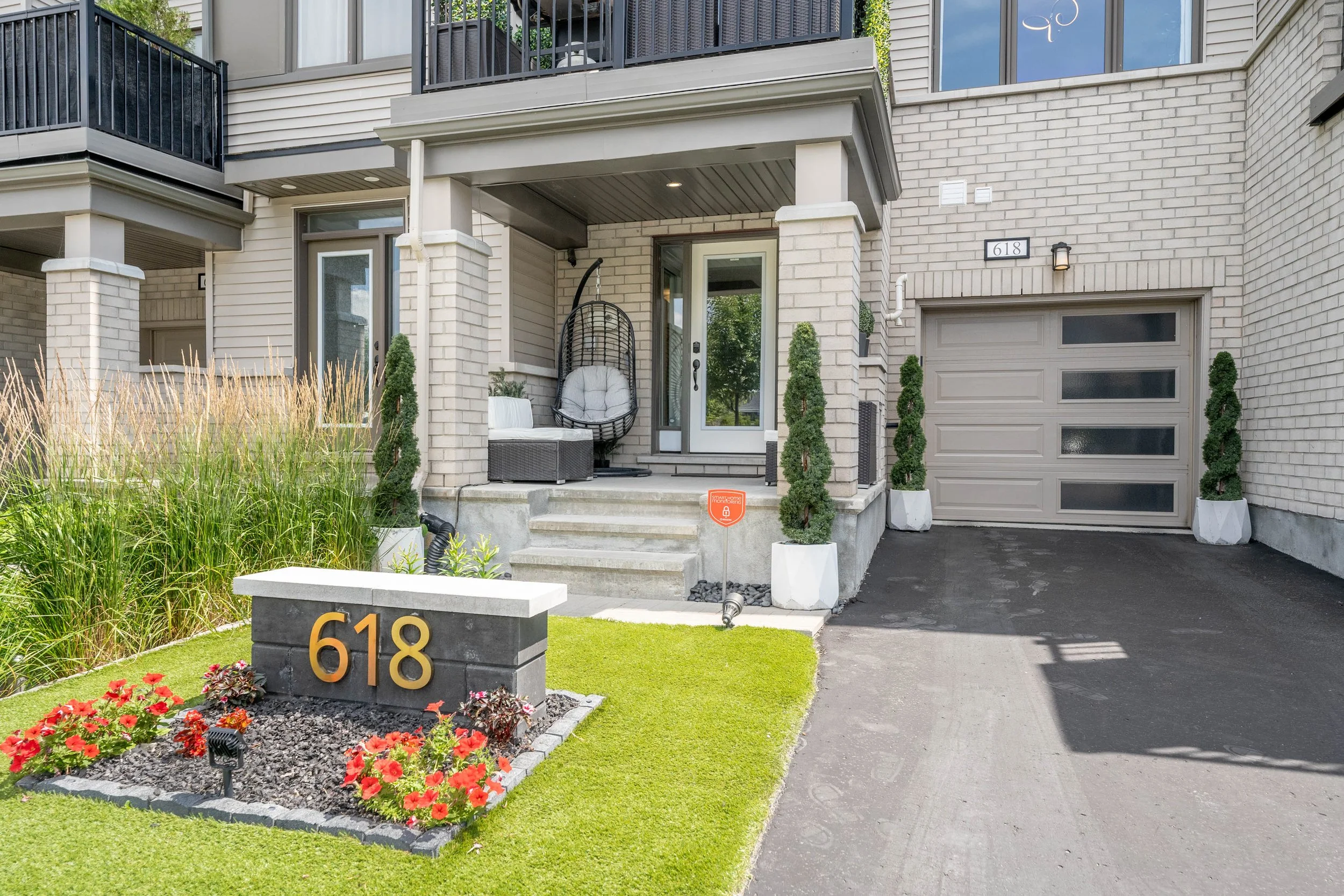 Front view of a modern townhouse with a small porch, potted plants, a hanging egg chair, and a well-maintained lawn with flower bed. The house number "618" is displayed on a stone sign and on the garage door.