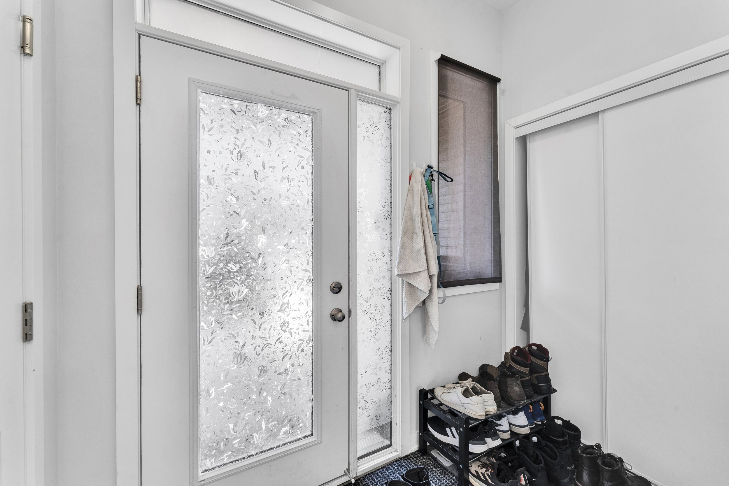 NV3D Real Estate Photography Ottawa  Entryway with a white door featuring decorative frosted glass, a shoe rack filled with various shoes, a wall hook with towels hanging, and a small window with a dark roller shade.