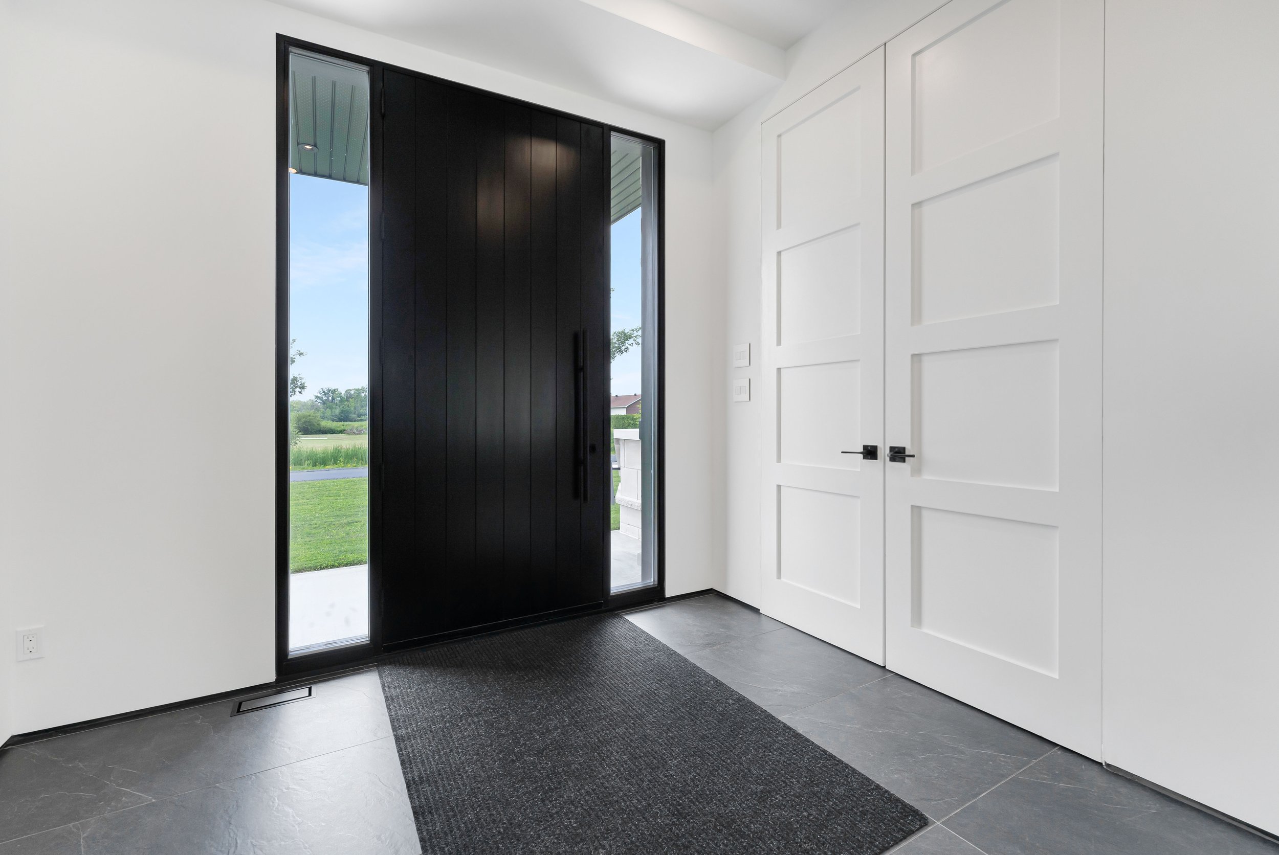 Modern entryway with black front door, white paneled walls, large window view, and slate tile floor.