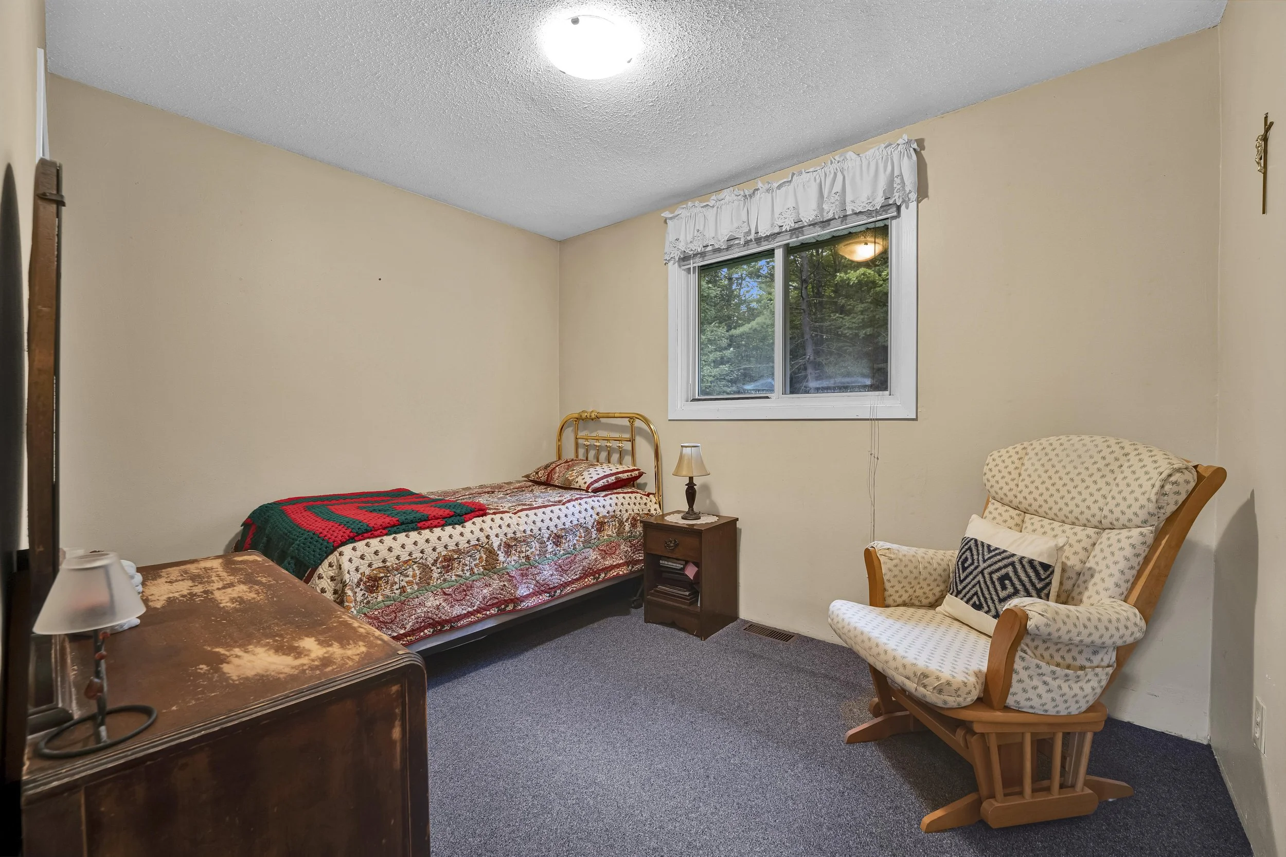 A bedroom with beige walls and a window showing trees outside. There is a bed with a colorful quilt, a small wooden nightstand with a lamp, a patterned armchair, and a wooden rocking chair.