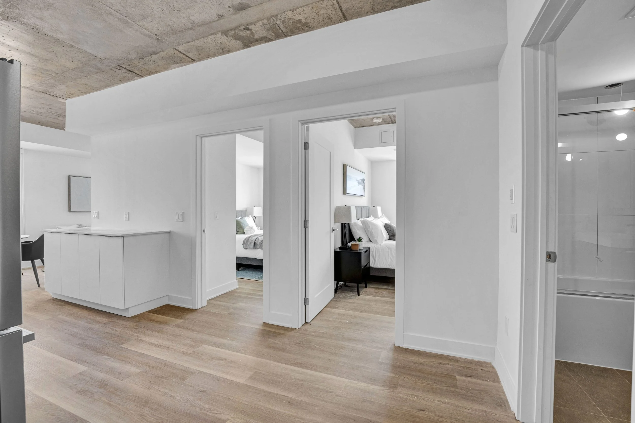 Ottawa Real Estate Media Services  Open doorway leading into a bedroom with a bed, nightstand, lamps, and wall art in a modern, minimalist interior with white walls and wooden floors.