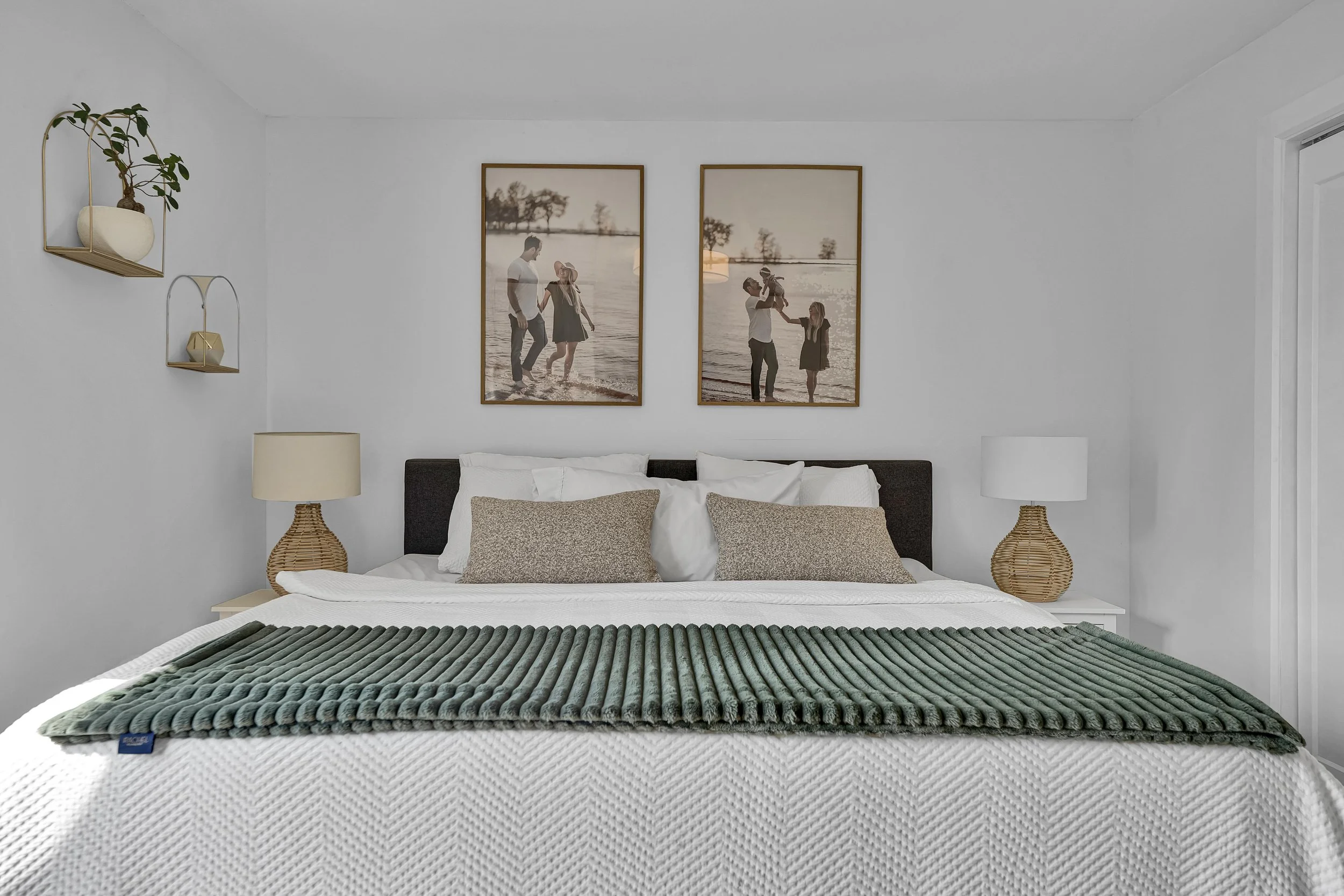 A modern bedroom with white walls and bedding, decorative pillows, two wicker bedside lamps, and framed photos of a family at the beach above the bed.