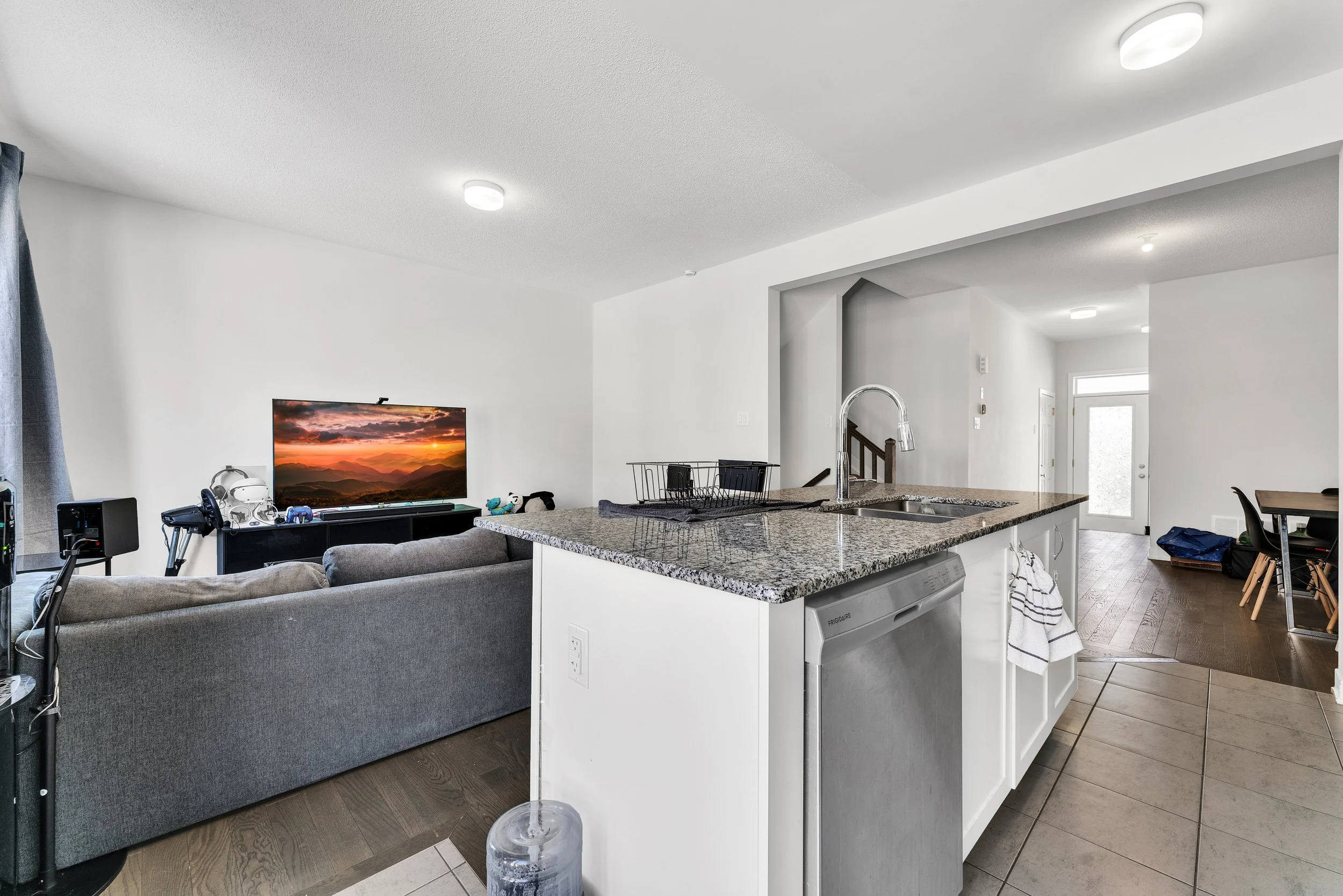 Open-concept living room and kitchen with gray sofa, TV displaying sunset, stone countertop with dish rack, and entryway in the background.