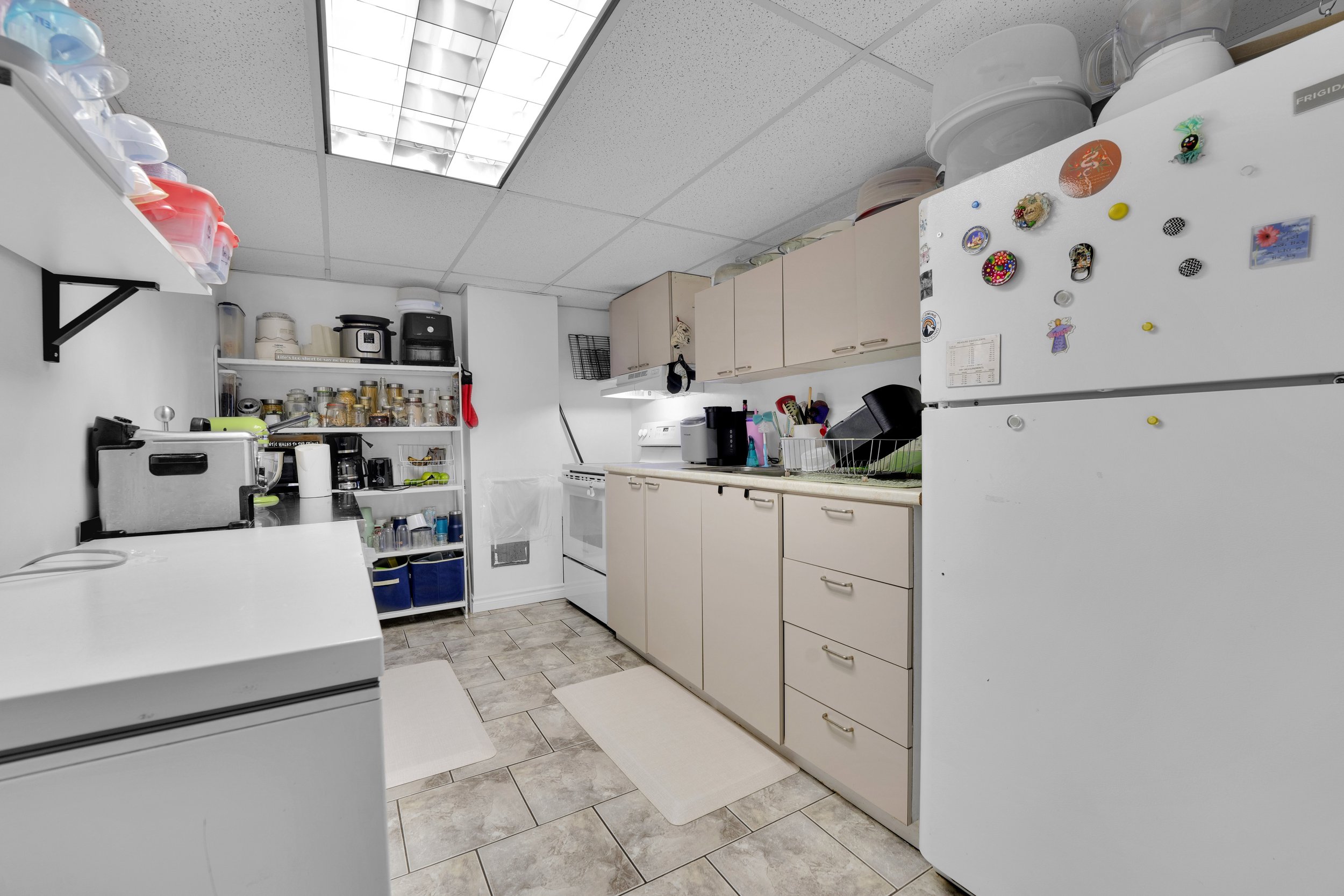 Ottawa Real Estate Media Services  A small kitchen with beige cabinets, white appliances, and gray tiled floors. There are various kitchen items, a freezer, and a microwave.