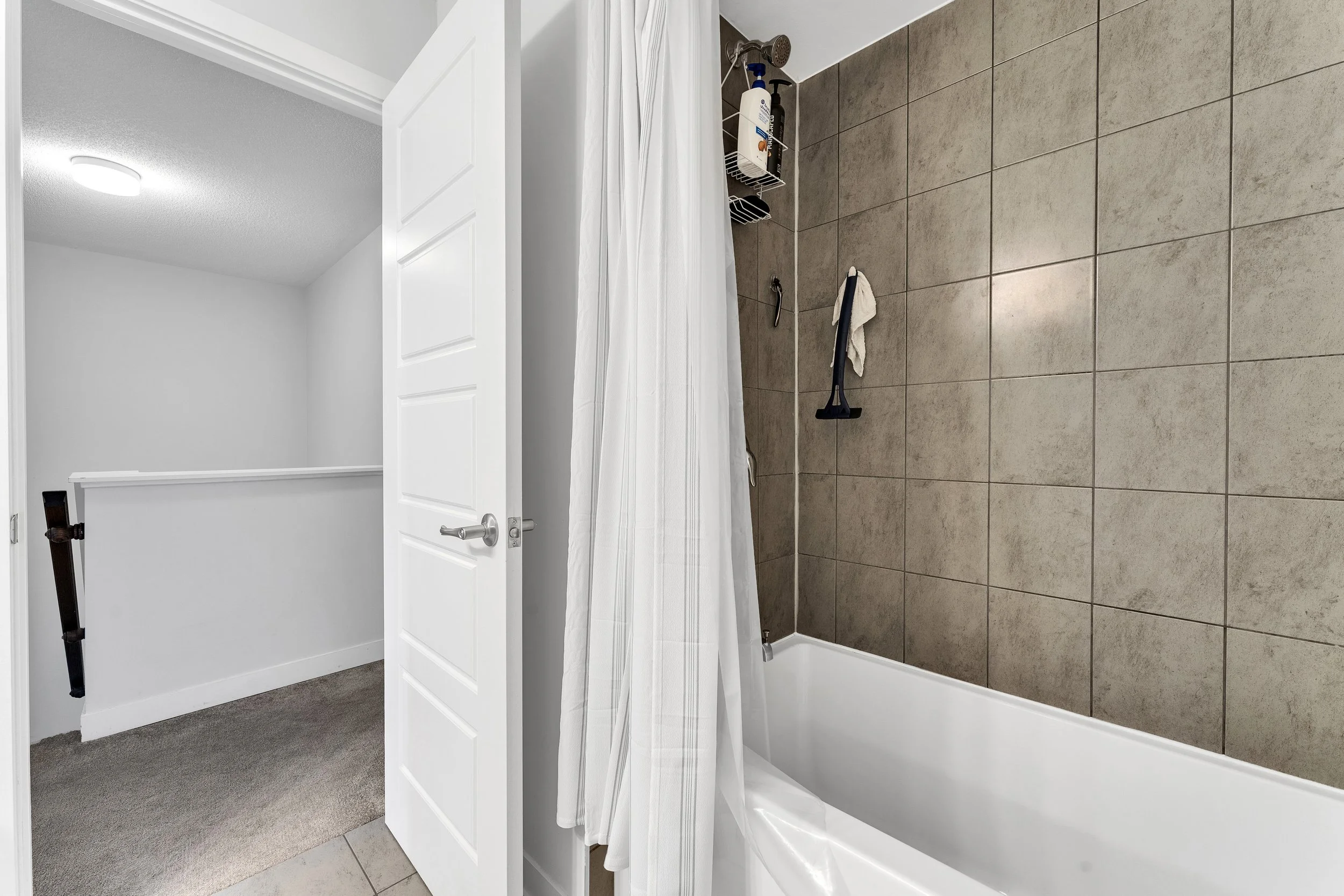 Bathroom with tiled shower, white bathtub, shower caddy, and white curtain. Adjacent room has white walls, door, carpeted floor, and a ramp for accessibility.