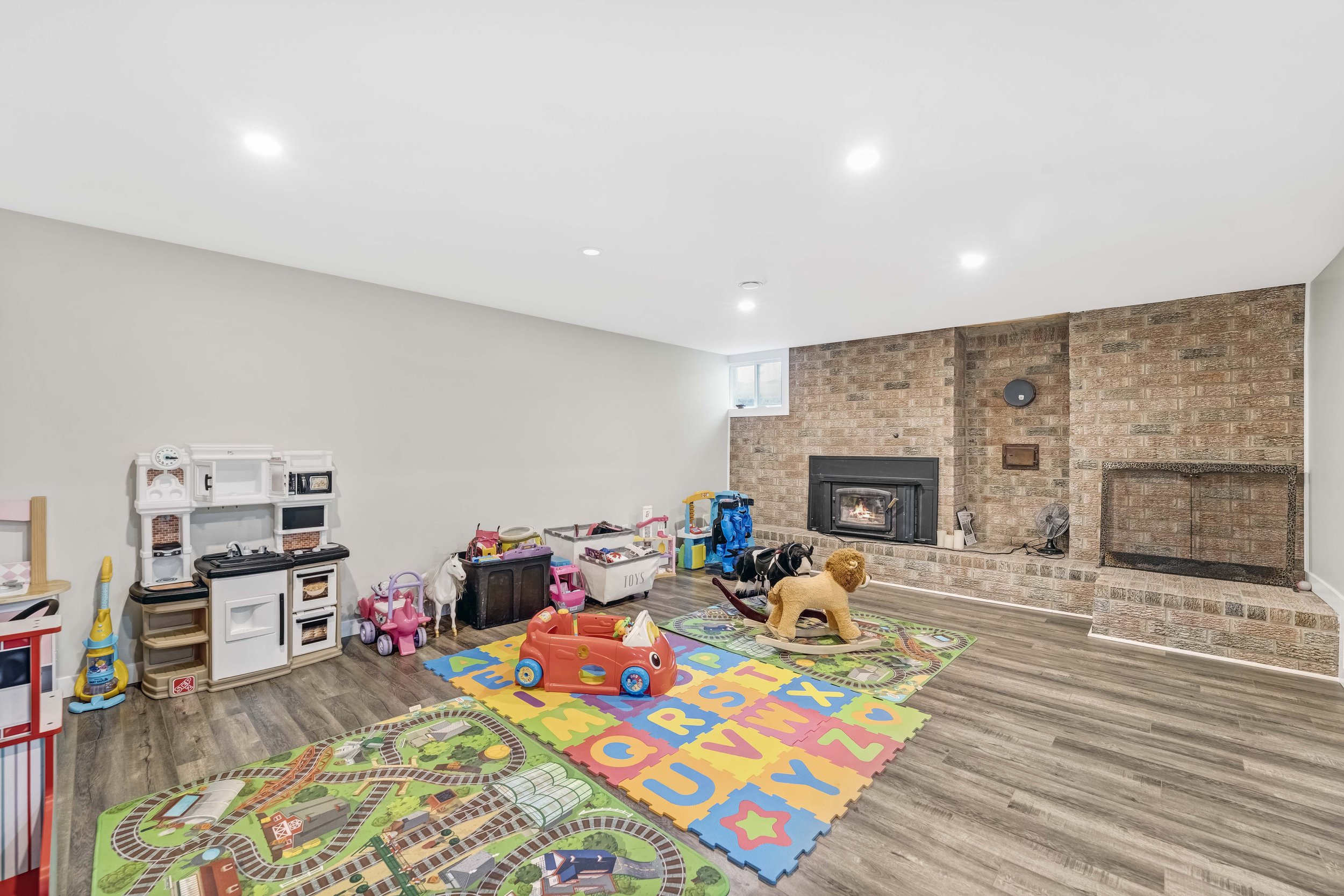 A playroom with colorful foam alphabet mats, a toy fireplace, and an assortment of children's toys near a brick fireplace with a fire burning inside, a small window in the corner, and a wooden floor.