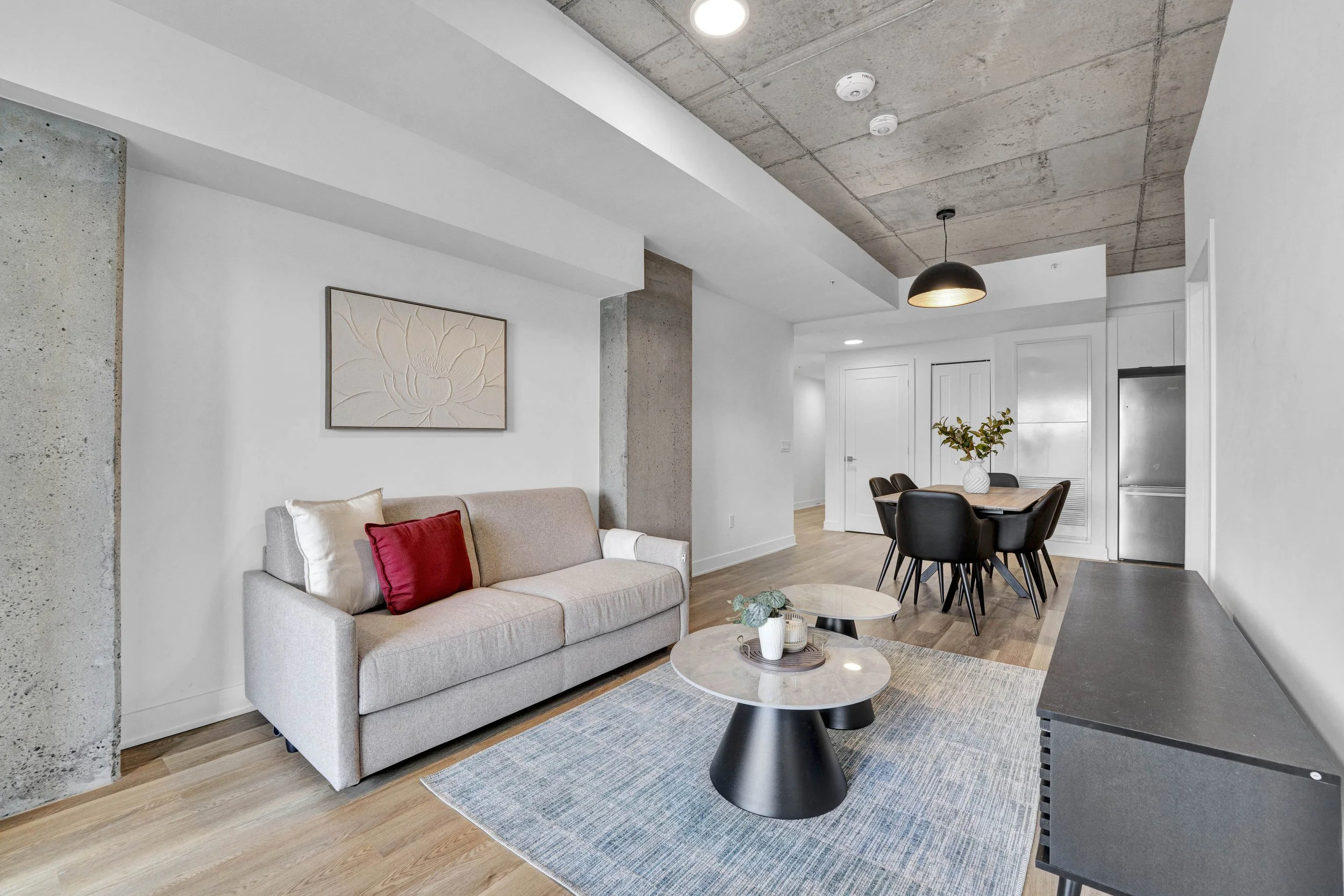 Modern living room with white walls, a beige sofa with red and white cushions, a blue area rug, round coffee table, and dining area with a table and black chairs. The ceiling features exposed concrete with recessed lighting and a black pendant light.