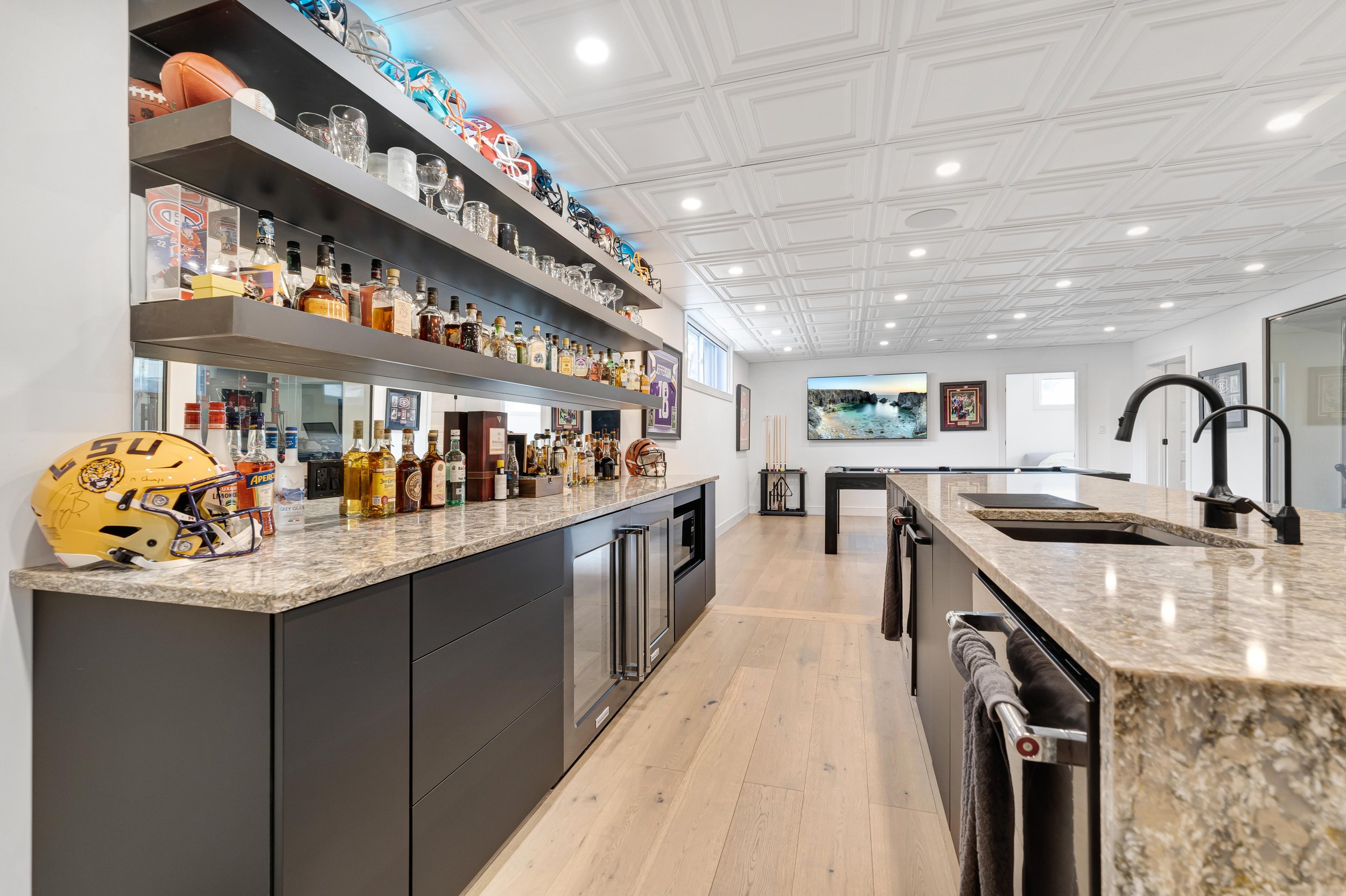 Modern home bar with shelves displaying liquor bottles and sports memorabilia, including football helmets and a signed LSU helmet, along with a foosball table and wall art in the background.