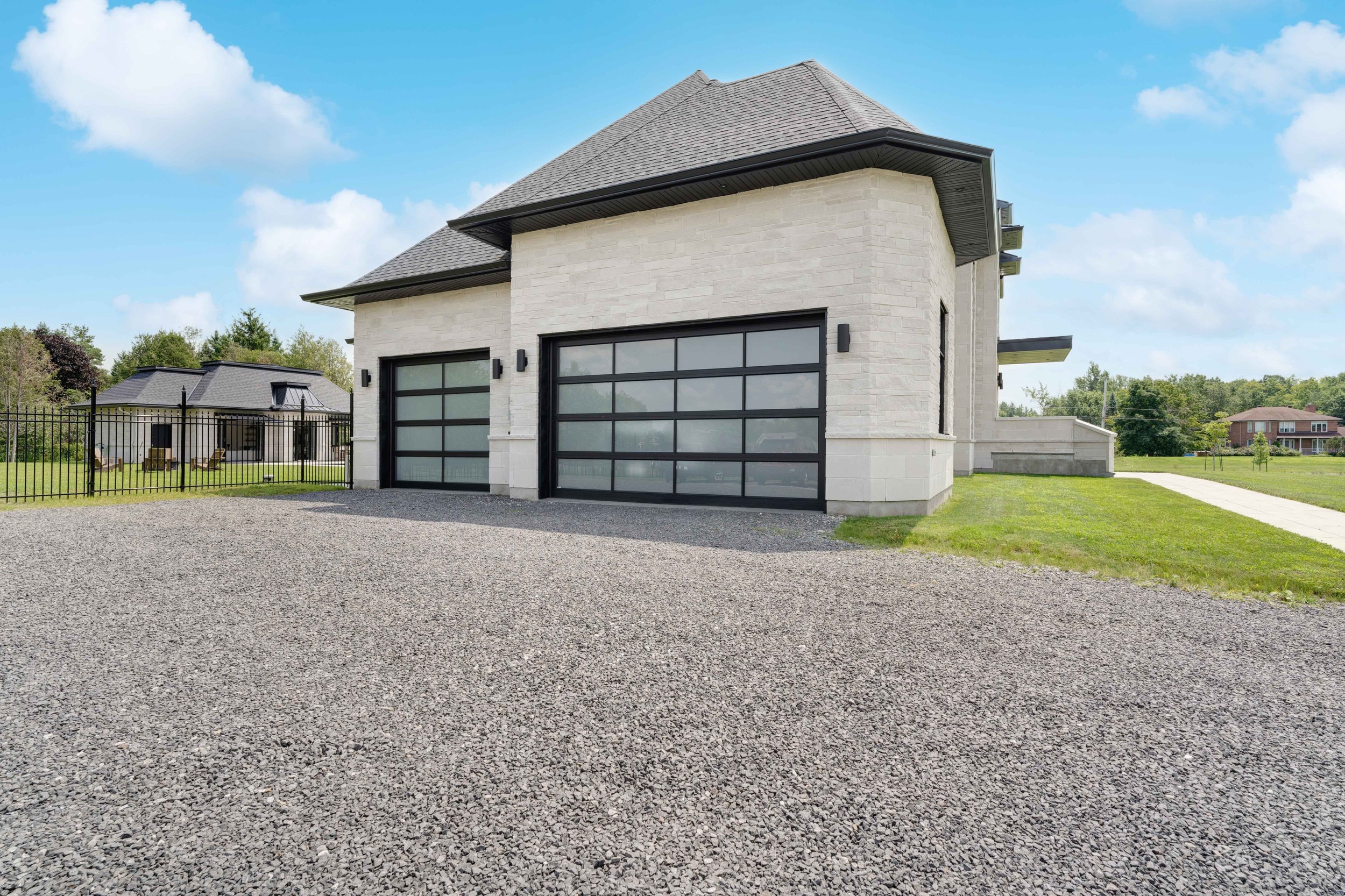 Modern stone house with large glass garage doors, paved driveway, and a detached smaller building in the background, surrounded by greenery under a blue sky.