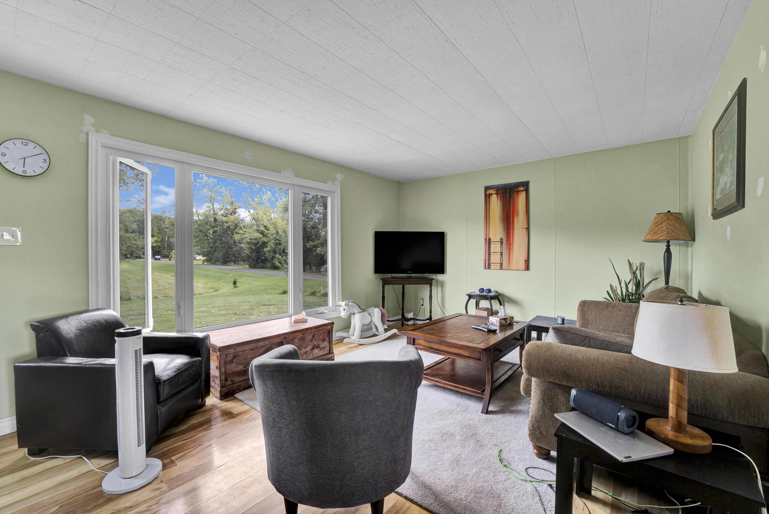 A living room with green walls and a large window showing a view of trees and a road outside. The room contains a black leather chair, a rocking horse, a flat-screen TV on a stand, a beige sofa, a wooden coffee table, a tall lamp, and wall art. There
