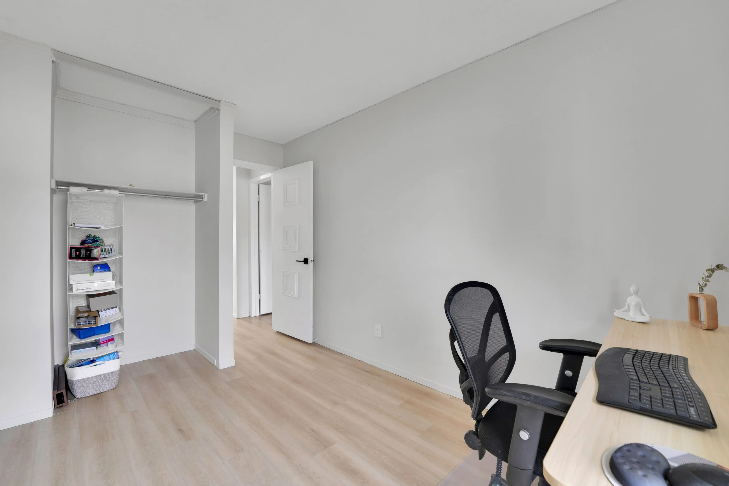A minimally decorated home office with white walls and light wood flooring. There is an open door leading to another room. On the left, a white storage shelf holds miscellaneous items. A black office chair and a wooden desk with a computer keyboard a