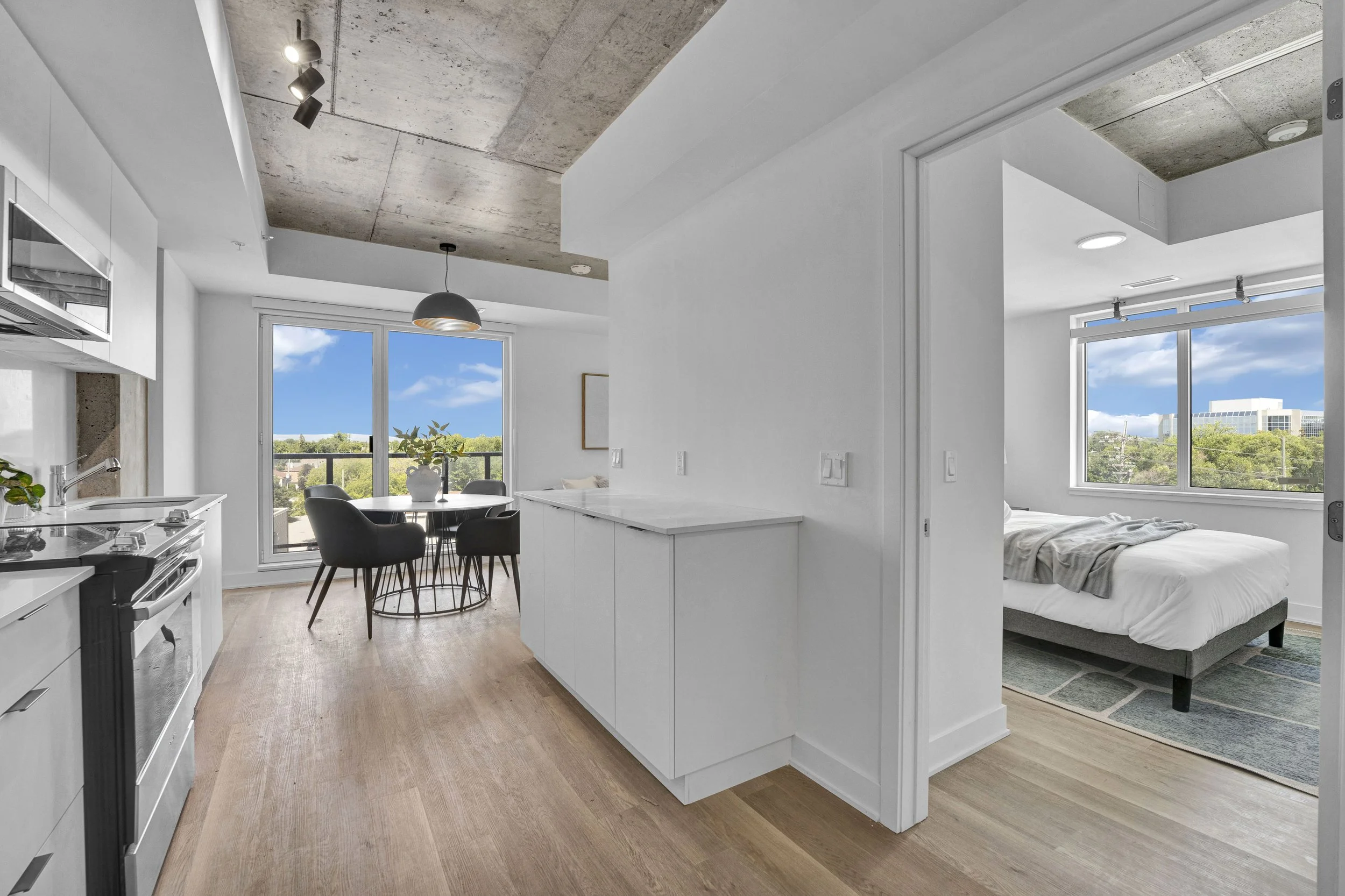 Modern apartment interior with kitchen, dining area, and bedroom, featuring white walls, large windows, and wooden flooring.