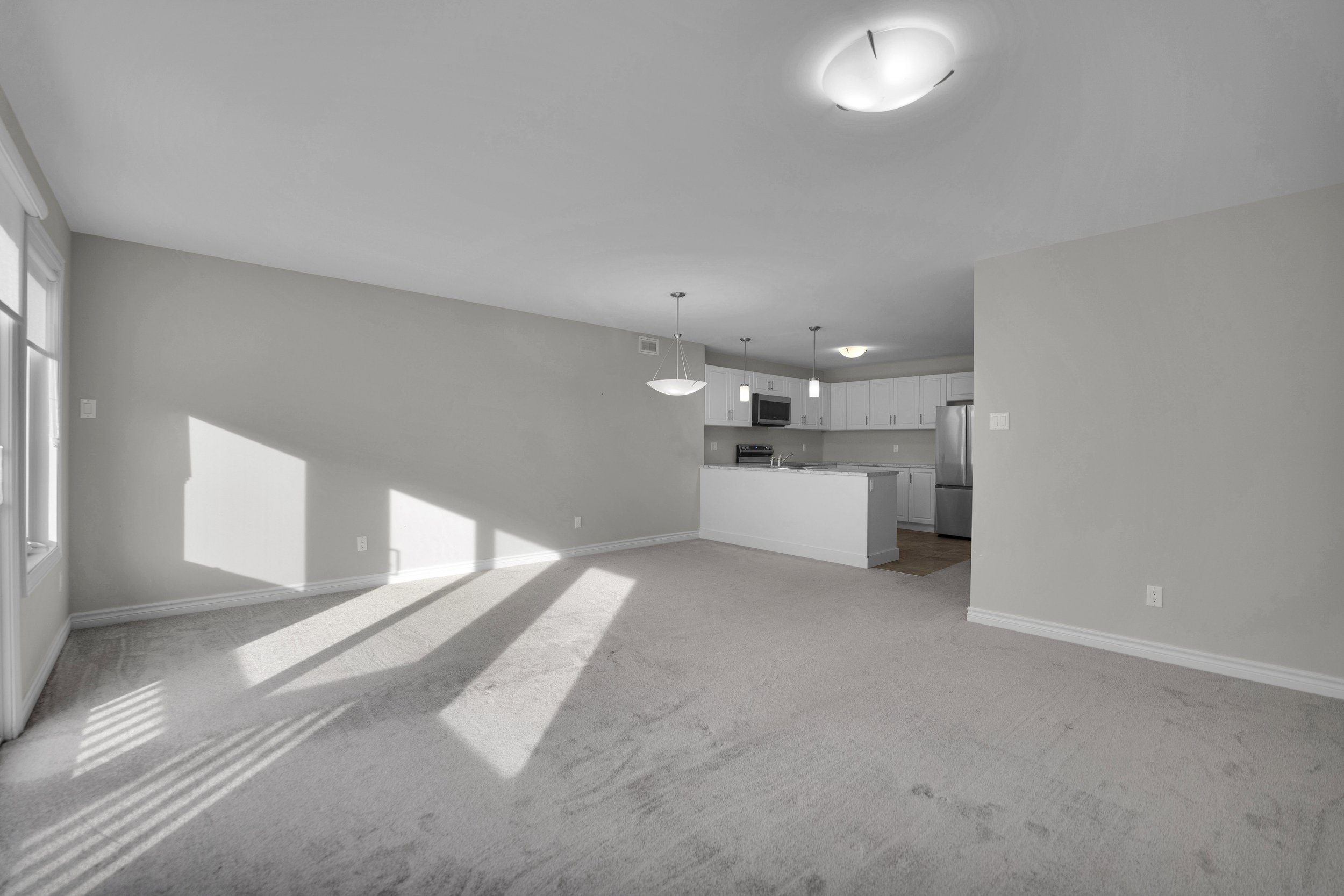 Empty living room with beige carpet, white walls, windows with blinds, and an open kitchen with white cabinets and stainless steel appliances.