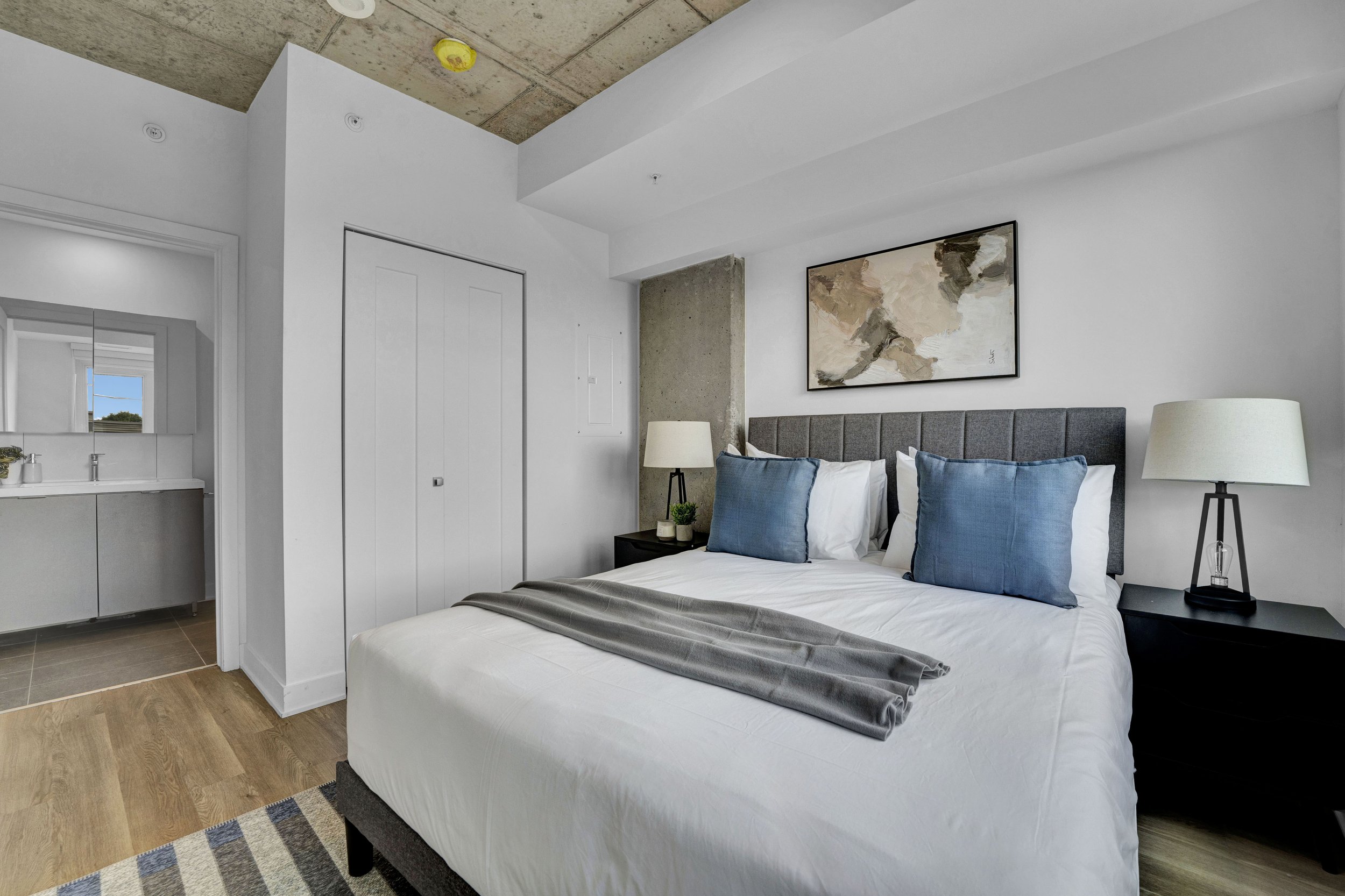 Modern bedroom with white bedding, blue pillows, black nightstands, lamps, abstract wall art, and a concrete ceiling with a smoke detector.