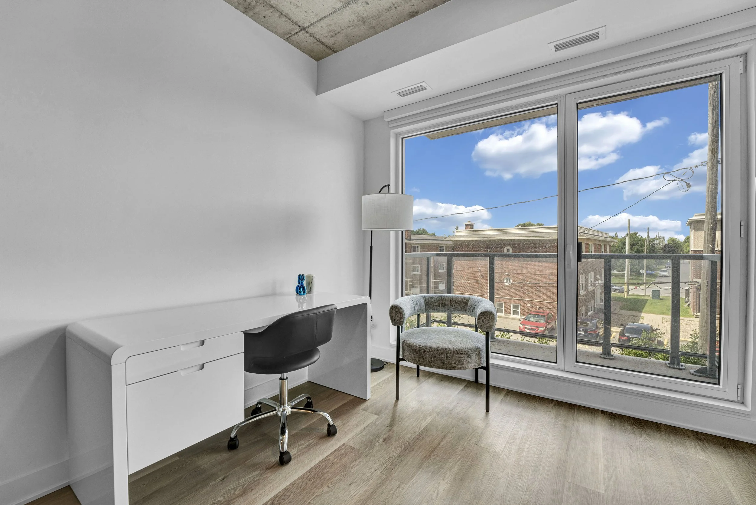Ottawa Real Estate NVISION 3D  Interior of a room with a large window showing a blue sky and buildings outside. Inside, there is a white desk, a black office chair, a round upholstered chair, a floor lamp, and some decorative items on the desk.