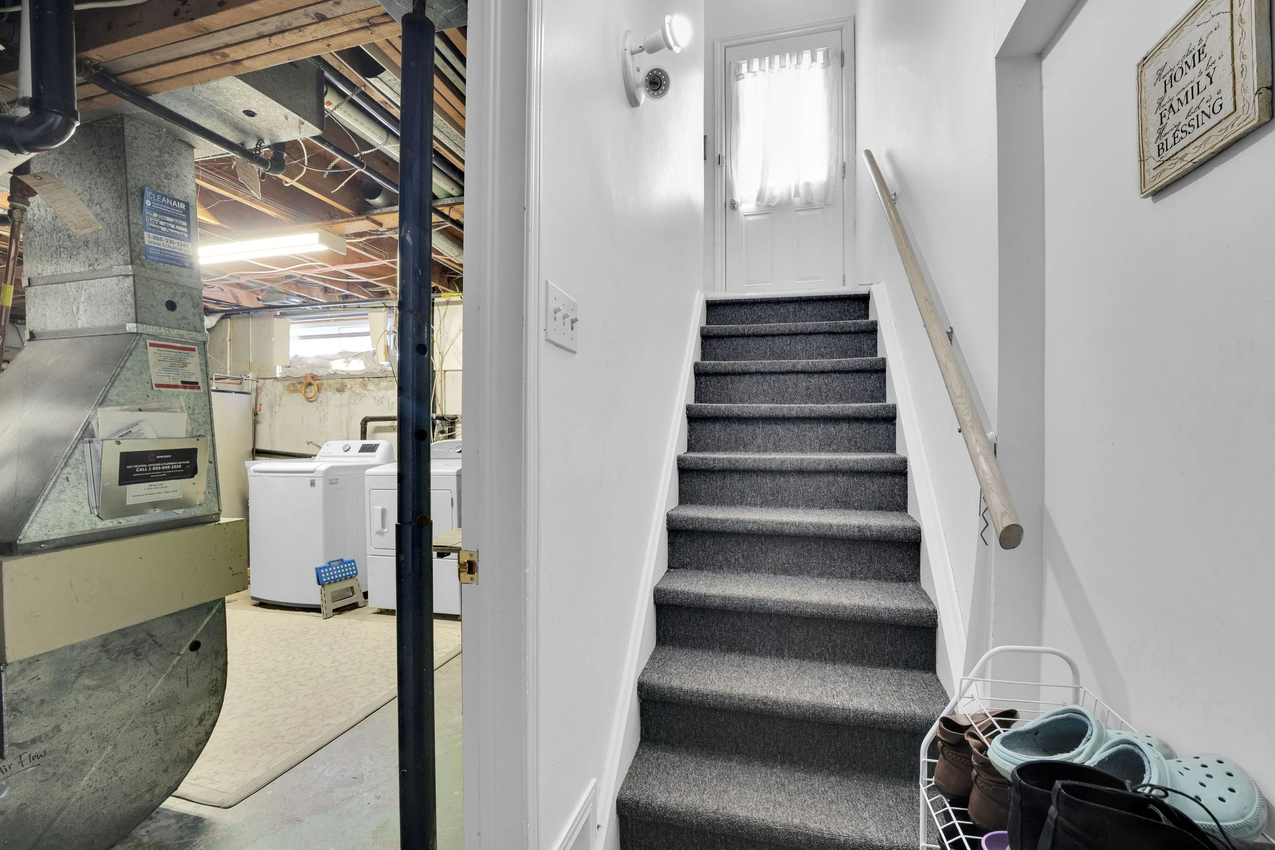 Interior view of a house showing a staircase with dark gray carpet leading upstairs, a wall-mounted light fixture, a white door, and a framed decorative sign that reads "Home, family, blessing." To the right, there is a small white shoe rack with foo