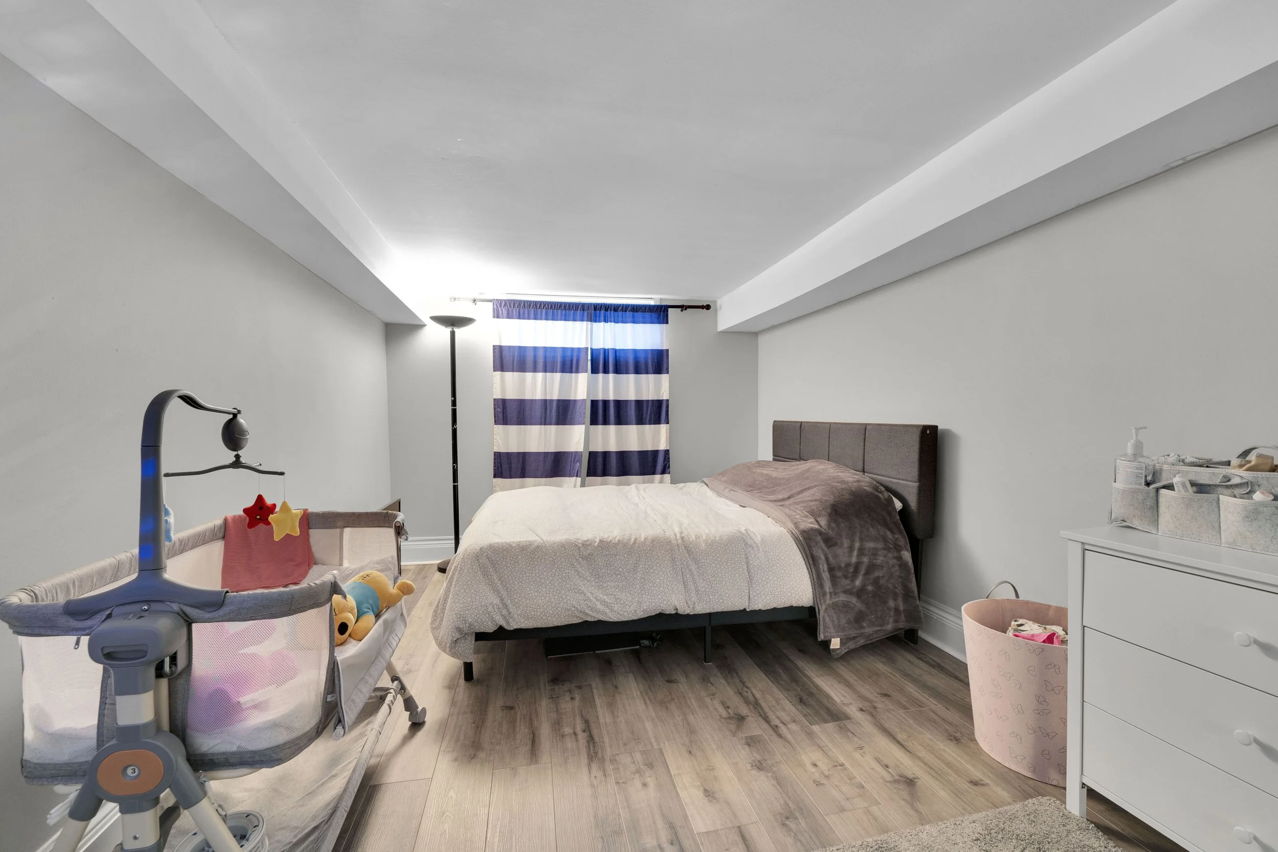 Ottawa Real Estate Media Services  A cozy bedroom with a bed, a window with blue and white striped curtains, a lamp, a dresser, and a crib with stuffed toys.