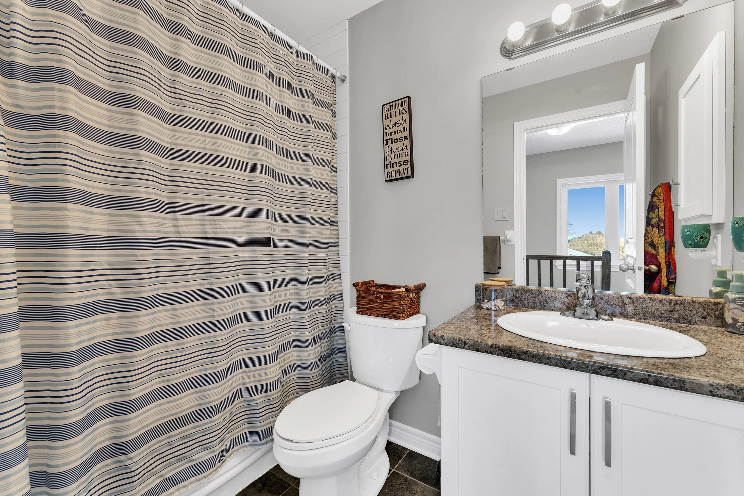 A bathroom with a striped shower curtain, a white toilet with a wicker basket on top, a granite countertop with a sink, a mirror above the sink, and a wall-mounted light fixture. A sign with bathroom rules is on the wall next to the mirror, and an op