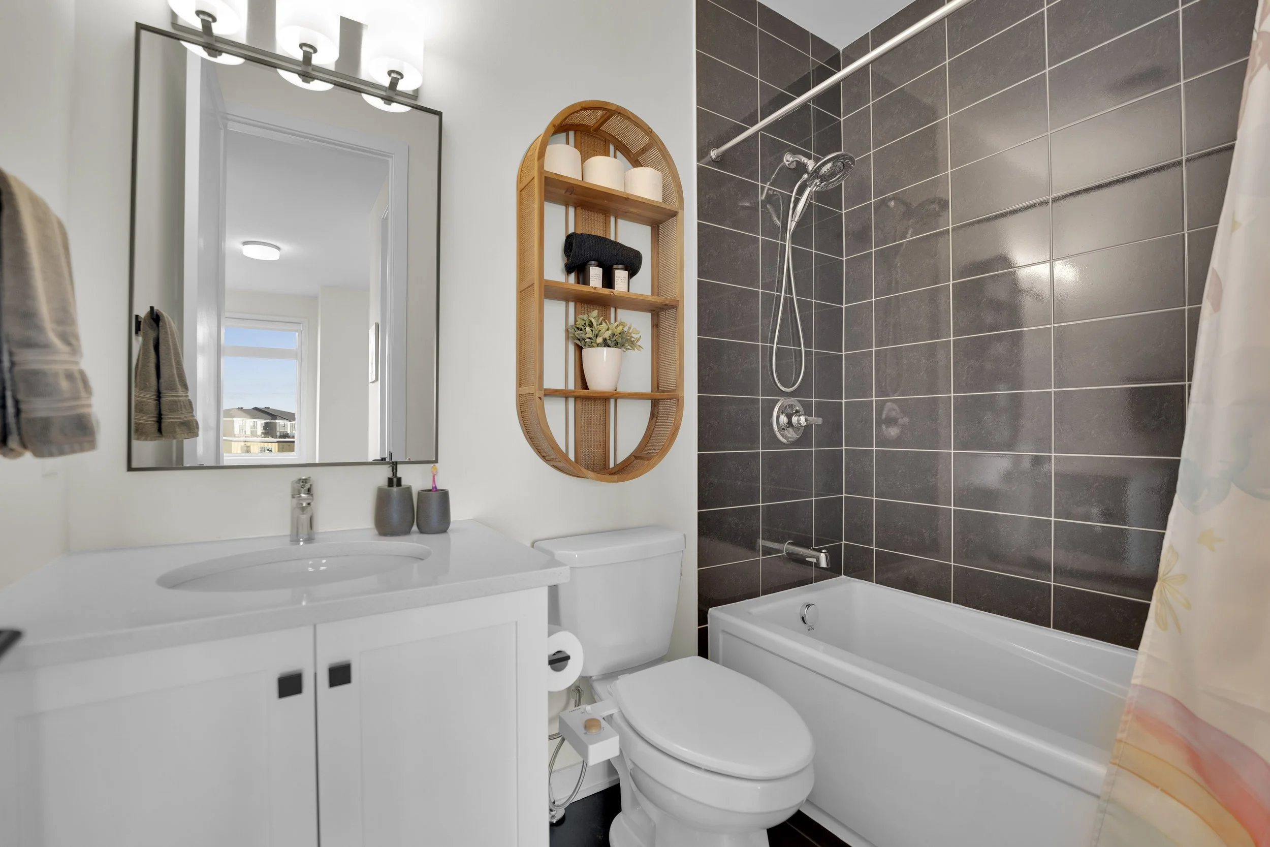 A compact bathroom featuring a white vanity with a built-in sink, a large mirror, and a gray soap dispenser. Above the mirror, there is a modern light fixture with three bulbs. Next to the vanity, a toilet with a bidet attachment is installed. To the