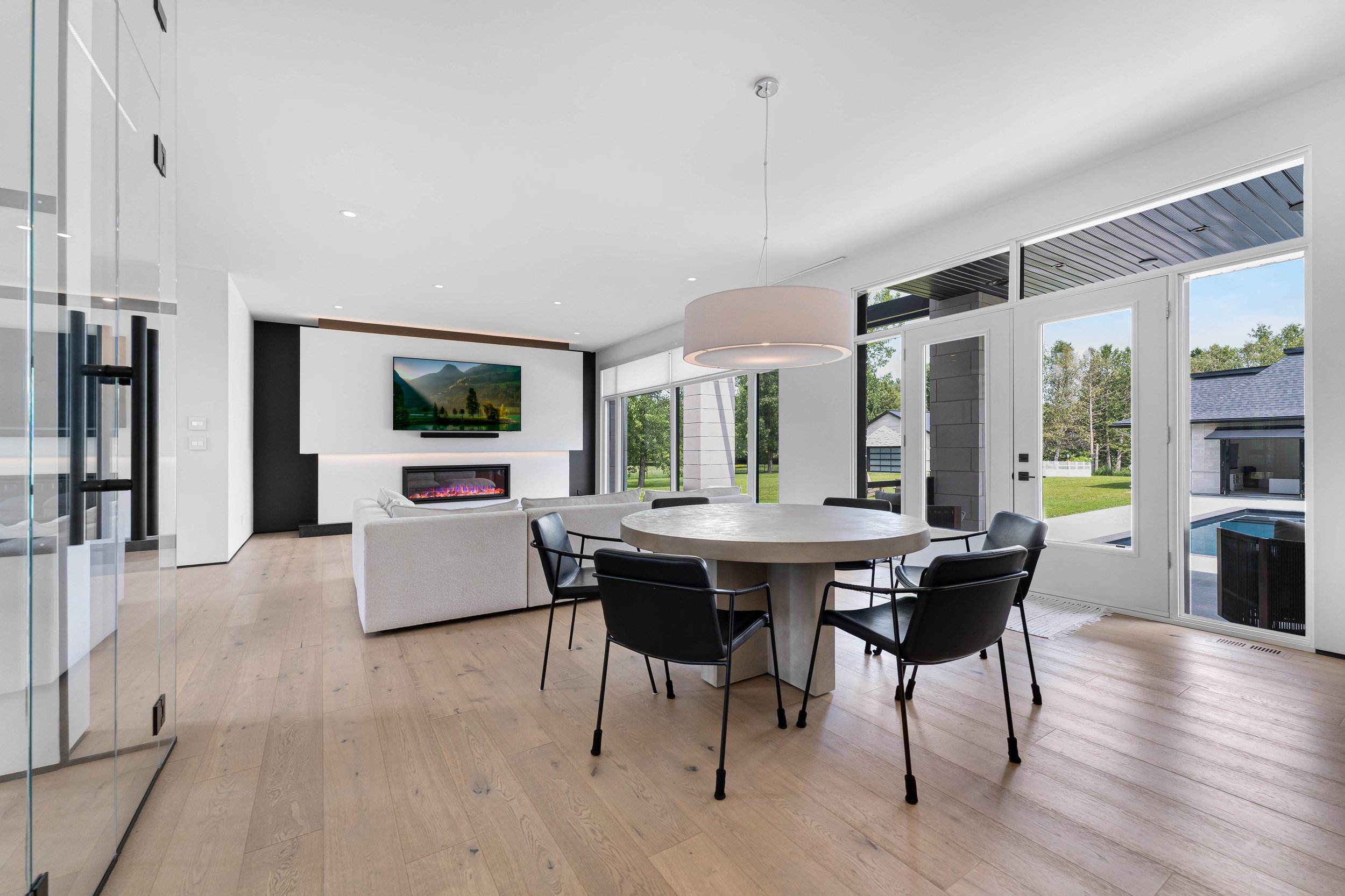Modern living room with wooden floors, a round dining table, black chairs, a ceiling light fixture, and a wall-mounted TV above a fireplace. Glass doors lead to a backyard with a pool. Real estate photography ottawa