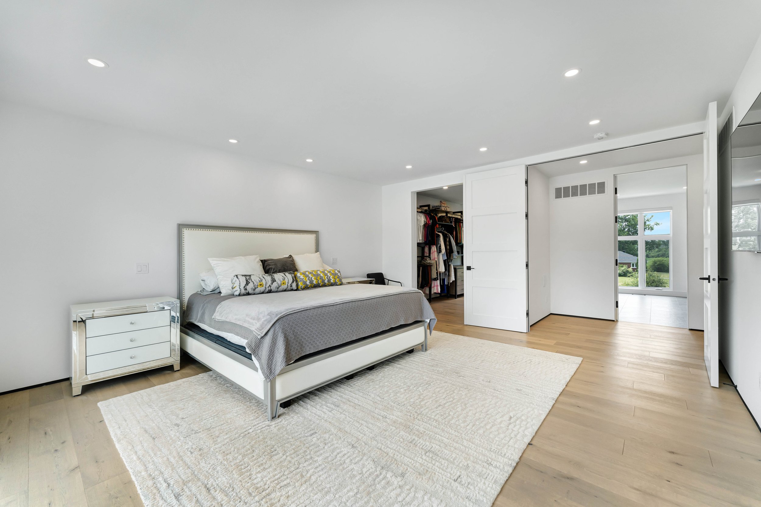 Modern bedroom with a large bed, white nightstand, open walk-in closet, light wood flooring, and a large window with a view of greenery.