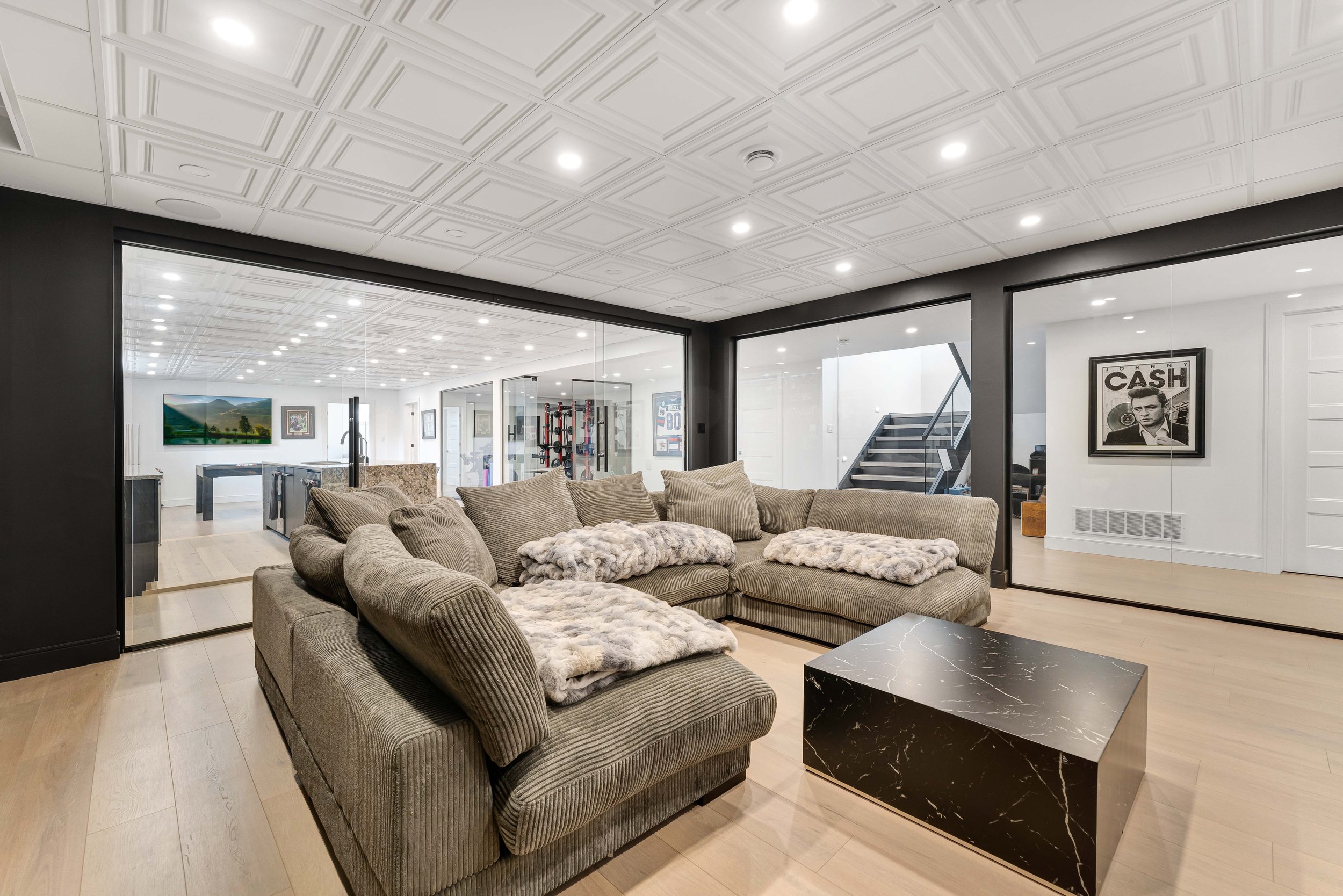 Modern living room with a large L-shaped sofa, marble coffee table, and decorative pillows. The room features a coffered ceiling and glass walls, leading to a recreational space with a foosball table. Art and framed pictures are displayed on the wall