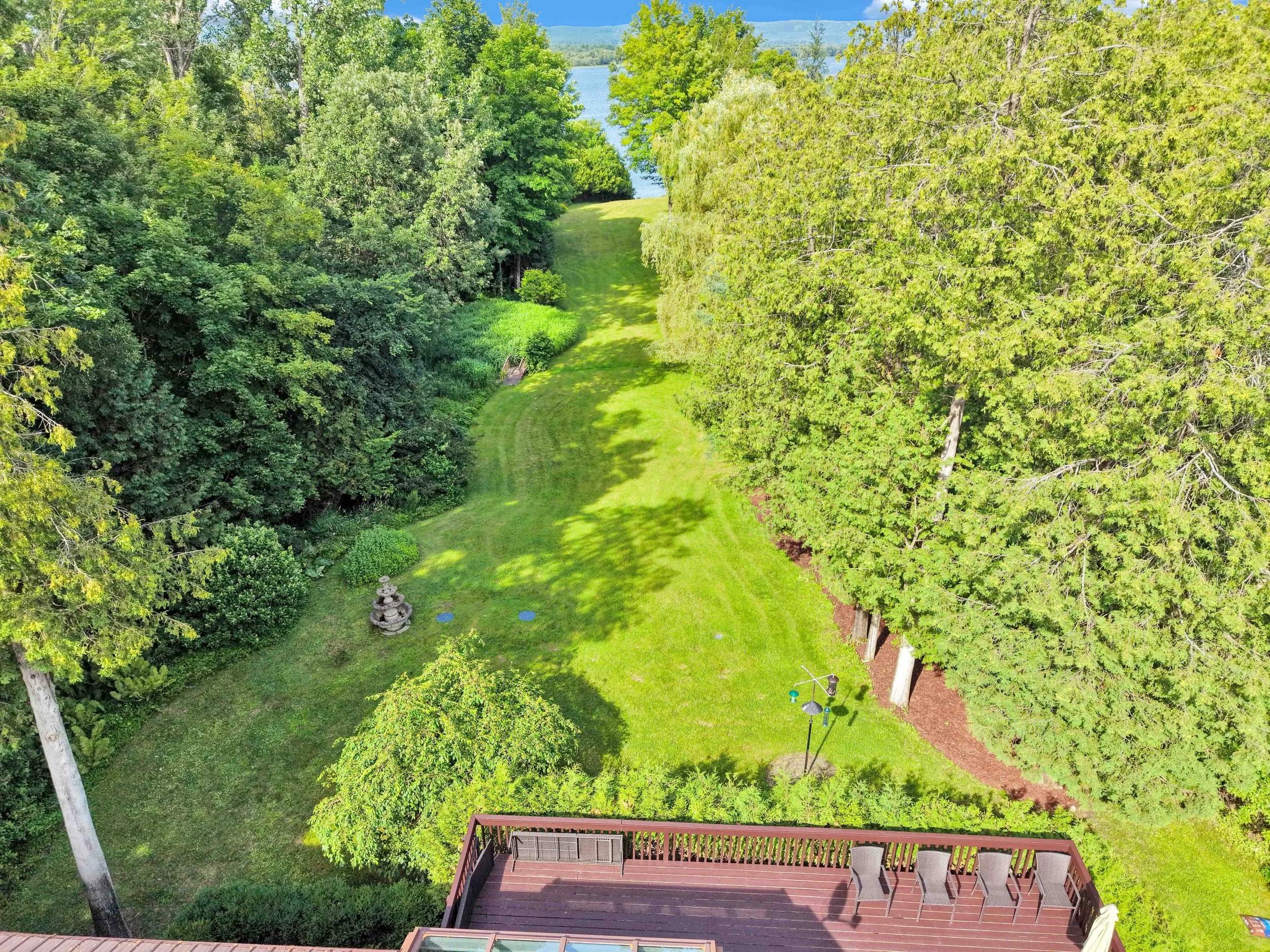 NVISION 3D Real Estate Media  Aerial view of a backyard with a grassy lawn, surrounded by tall green trees, and a deck with outdoor furniture. In the background, there is a lake and distant mountains under a blue sky.