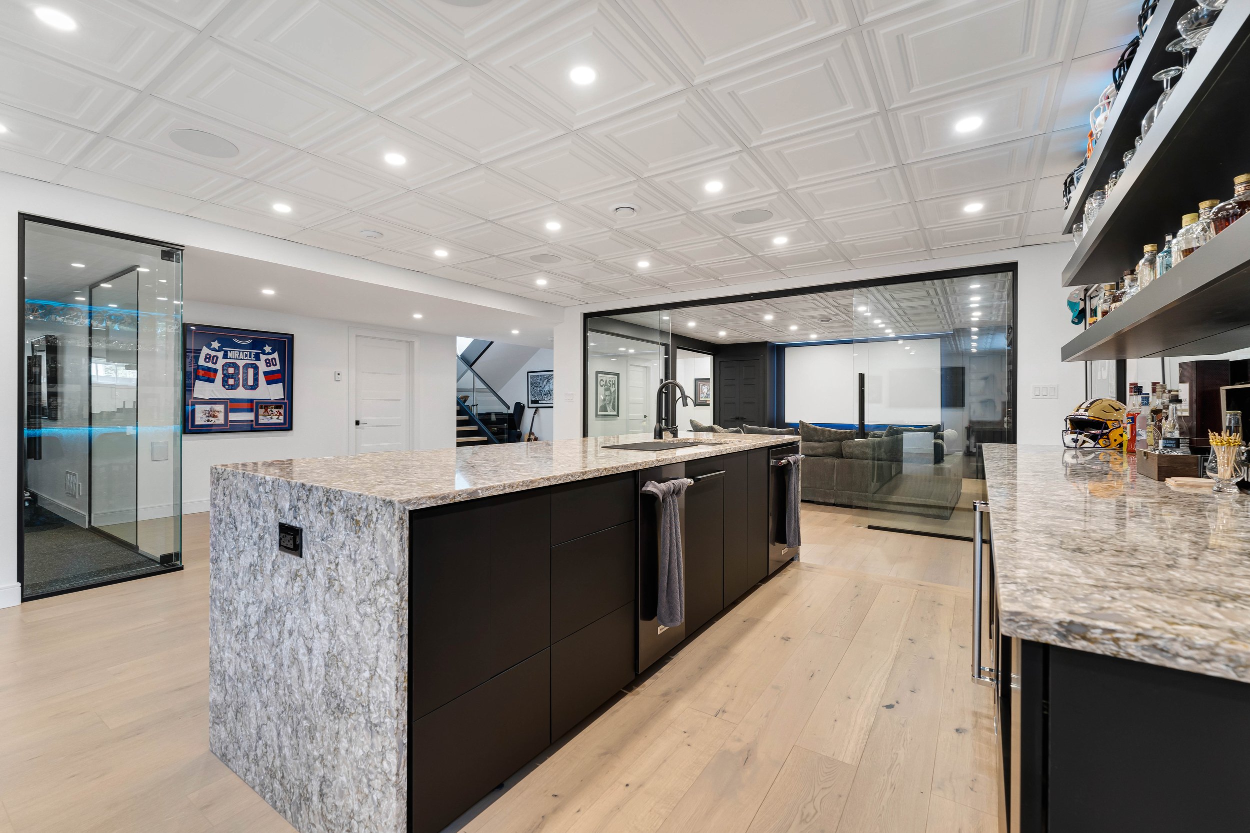 Modern basement with a kitchen island, bar area, and sports-themed decor, featuring a glass wall, framed jersey, and seating area.