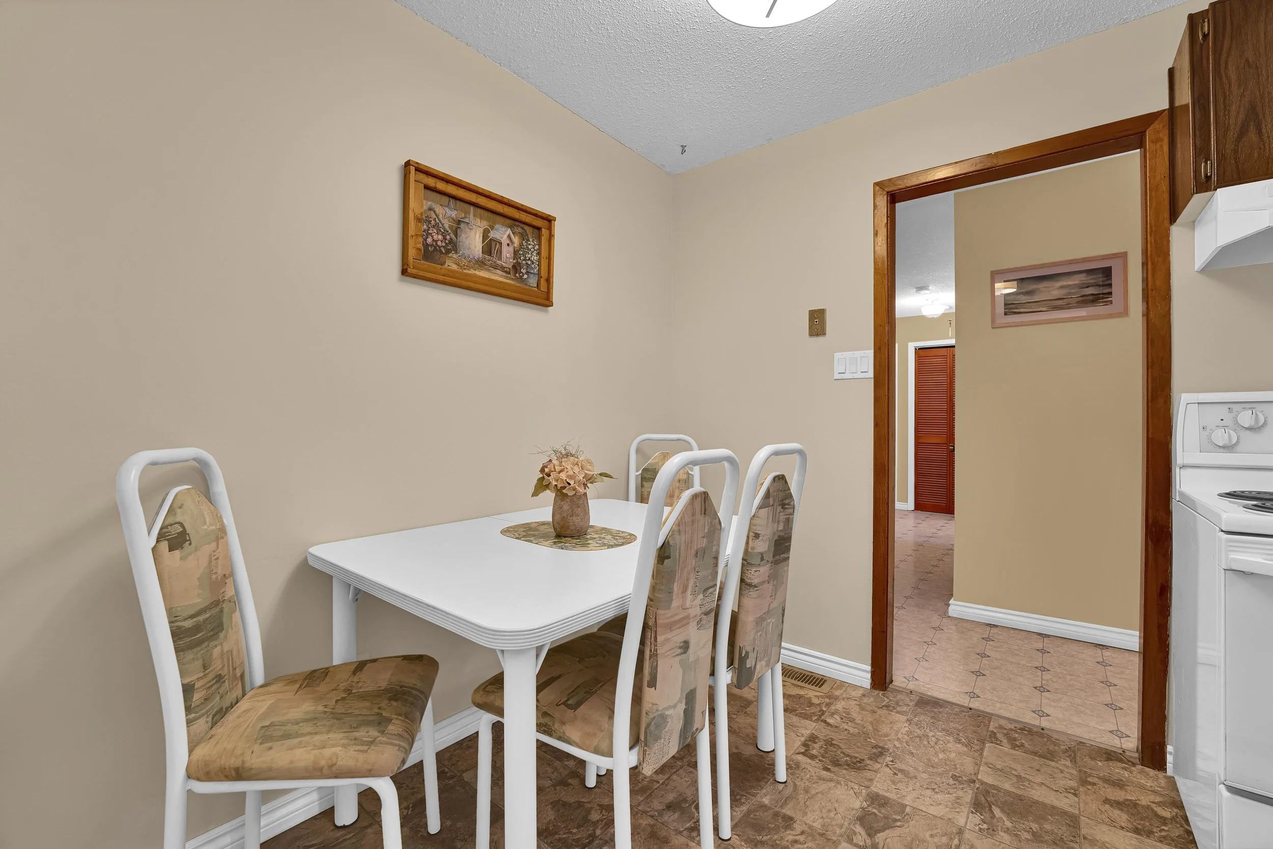 Ottawa Real Estate A dining area with a white table and six chairs with patterned fabric seats and backs, a small flower arrangement on the table, and artwork on the walls. The room leads into a kitchen with a stove and a doorway with a wooden frame.