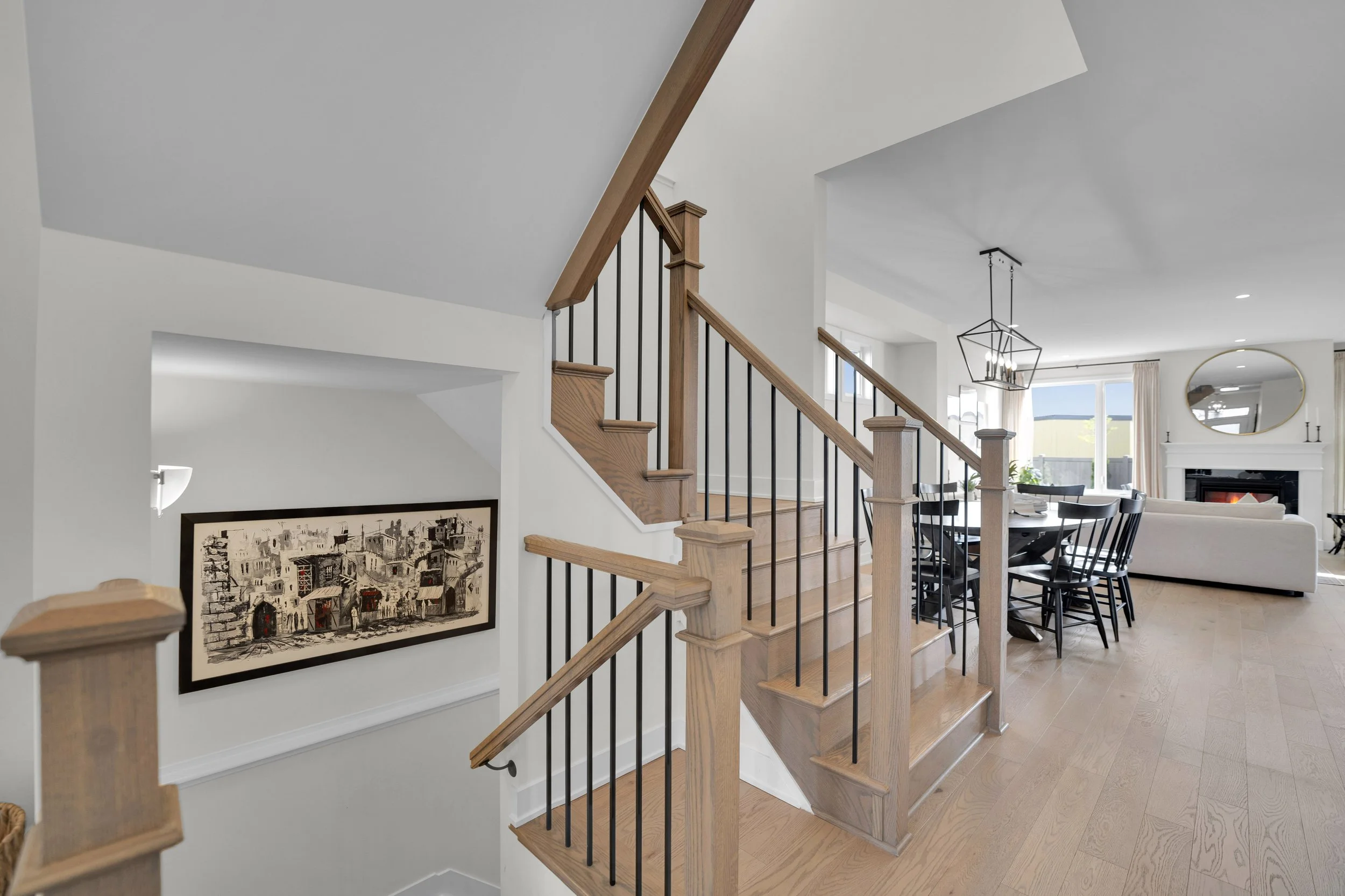 Interior view of a modern home featuring a staircase with wooden steps and black metal railings, leading to the upper floor. The open floor plan includes a dining area with a black dining table, black chairs, a living room with a white sofa, a firepl