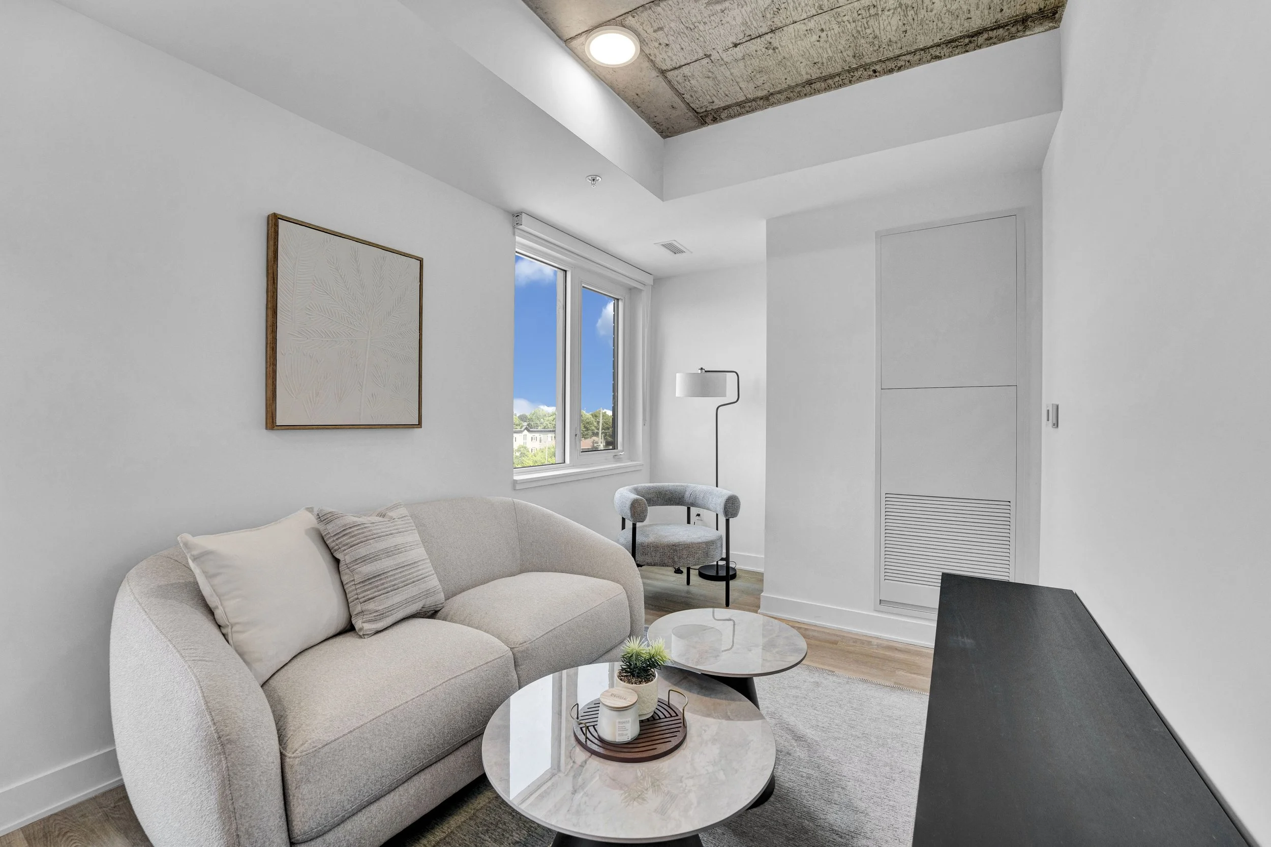 Interior living room with white walls, a beige sofa with pillows, a small round marble coffee table with decorative items, a gray armchair, a floor lamp, and a window showing blue sky and trees.