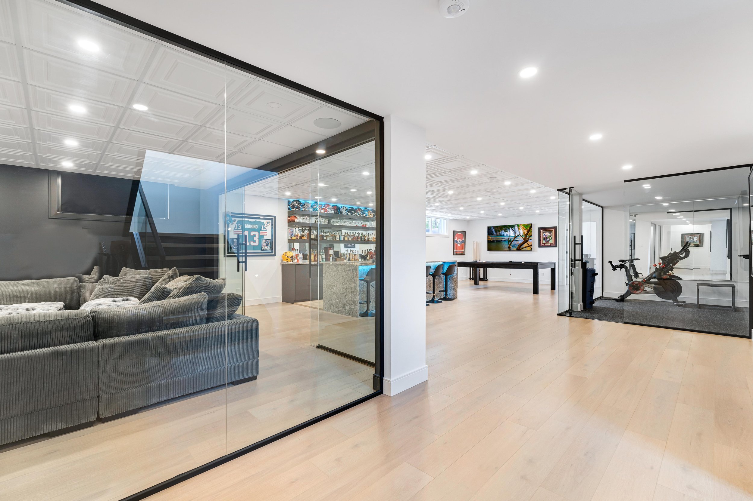 Modern basement with glass-enclosed sitting area, home bar, game room, exercise bike, and wall decor in a well-lit space.