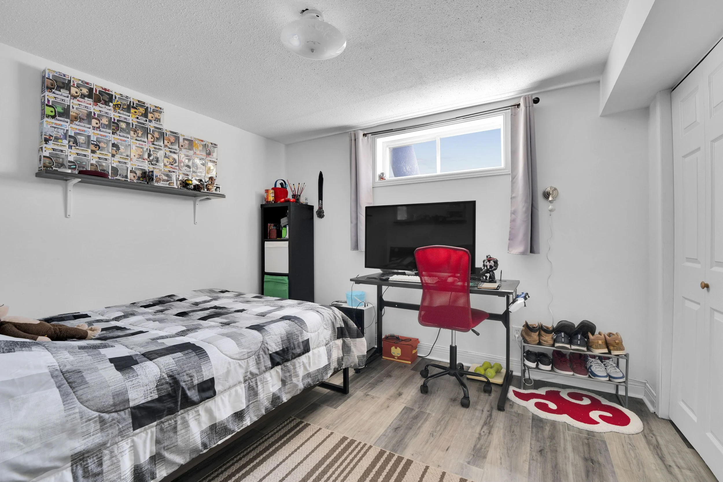 A bedroom with a bed featuring a black and white checkered comforter, a wall-mounted shelf with a collection of Funko Pop! figures, a black desk with a red office chair, a television, a black bookshelf, and shoes arranged on a shoe rack. There is a w