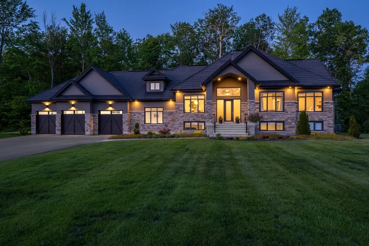 Luxury stone house with large windows, three-car garage, and well-manicured lawn, surrounded by trees at dusk.