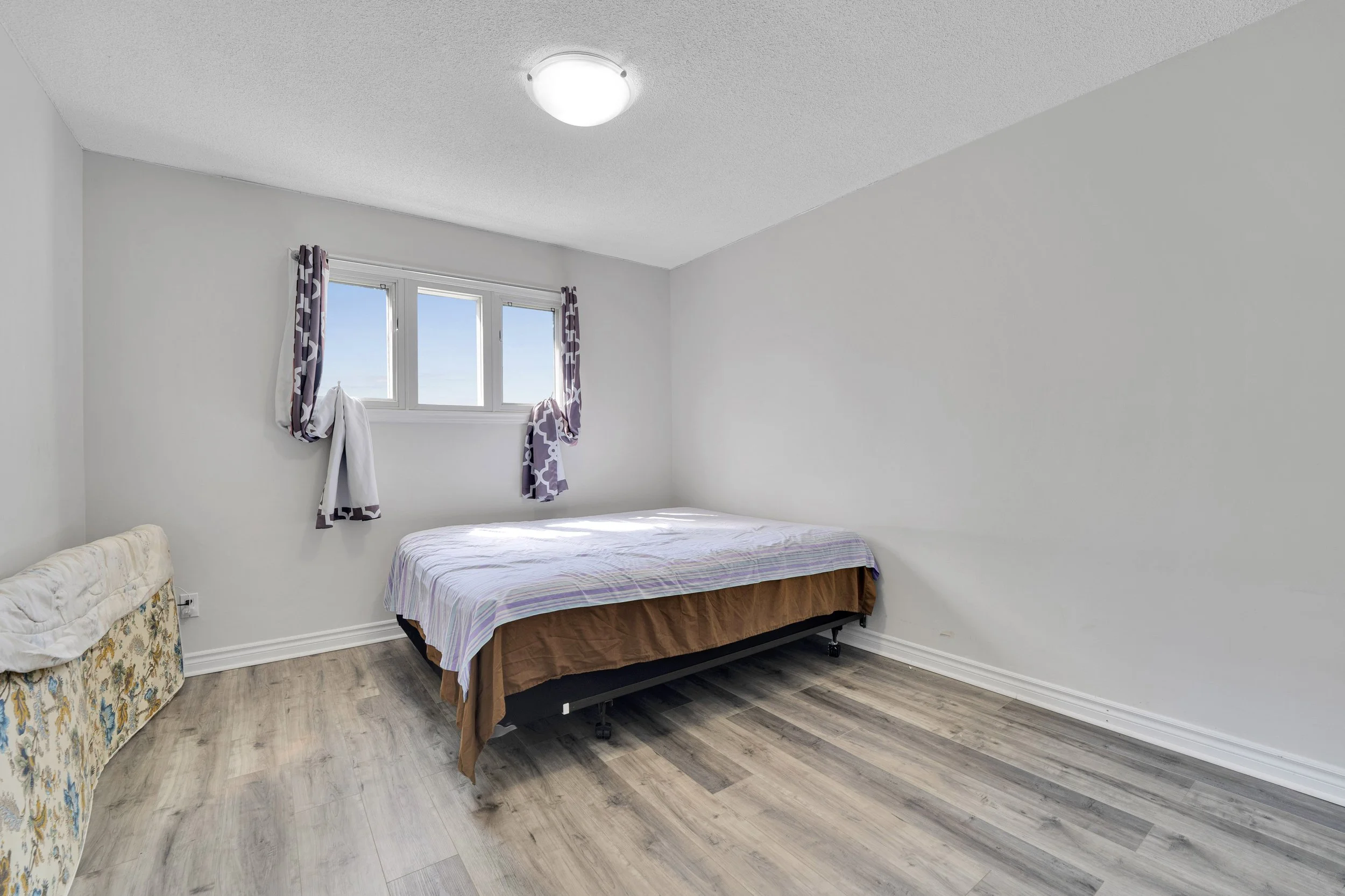 Ottawa Real Estate Media ServiA bedroom with light-colored walls and a wood-style floor. There is a window with patterned curtains, a filled bed with a striped lavender and white sheet, and an upholstered armchair with a floral pattern in the corner.