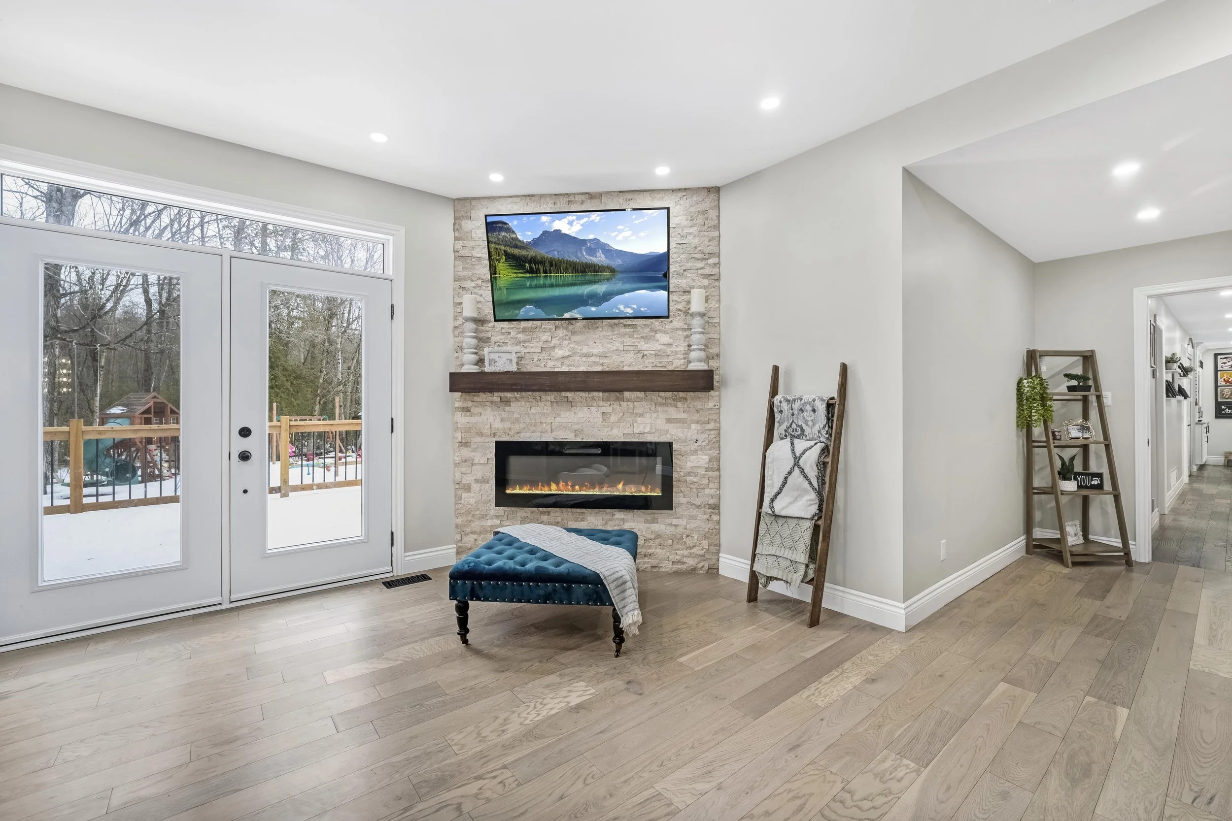 Living room with a stone fireplace, mounted TV, hardwood floors, glass doors leading outside to a snow-covered yard, and a blue tufted ottoman. Ottawa Real Estate Photography