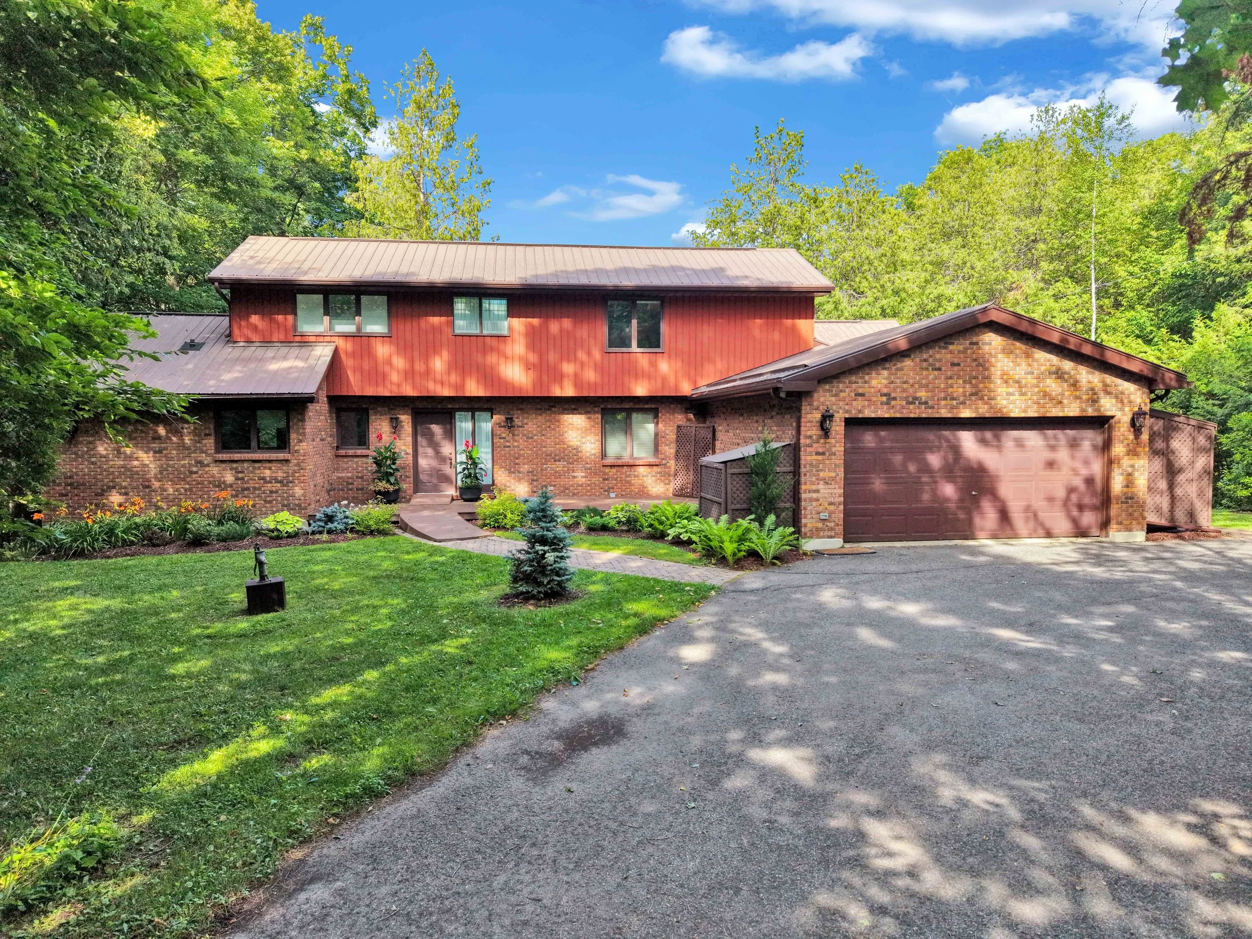 NVISION 3D Real Estate Media  A two-story house with red siding, brick lower level, and a brown metal roof, surrounded by trees and a well-maintained lawn with a curved driveway.