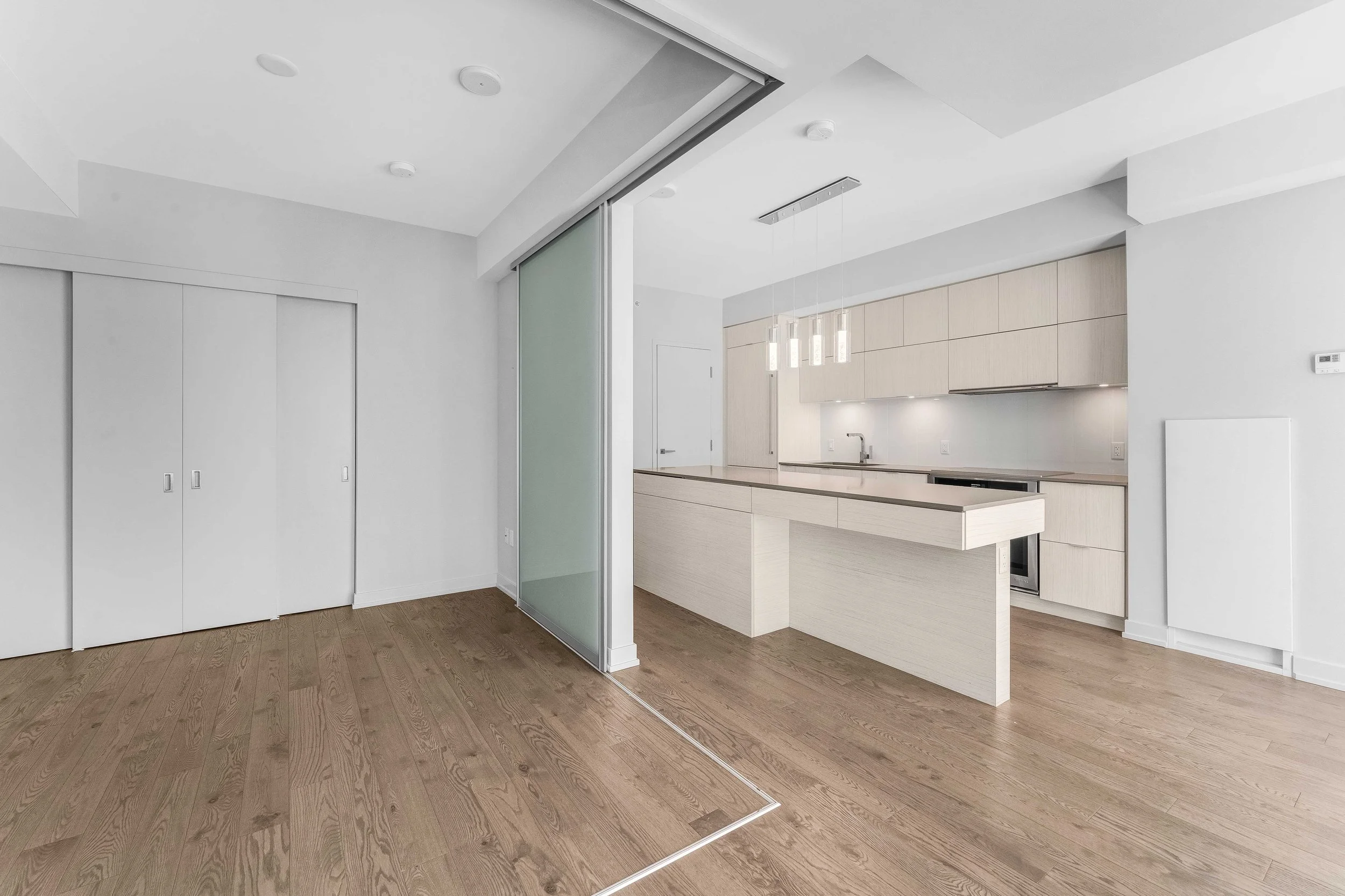 NV3D Ottawa Real Estate Photography Modern empty kitchen with light wood cabinets, a white countertop, minimalist pendant lights, and sliding glass door separating from living space with hardwood floors.
