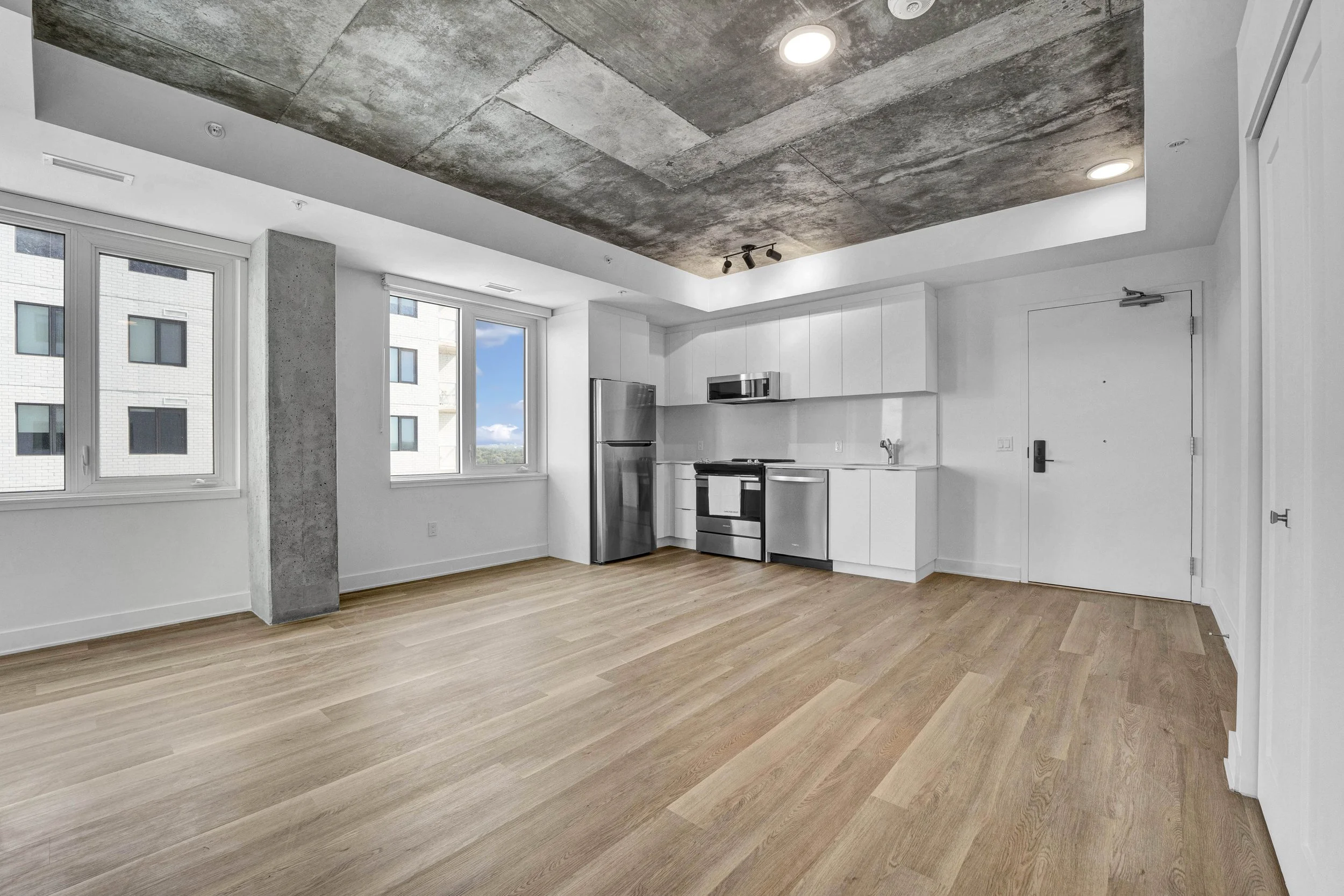 Empty modern kitchen with white cabinets, stainless steel appliances, wooden flooring, and a concrete ceiling with recessed lighting and track lights.