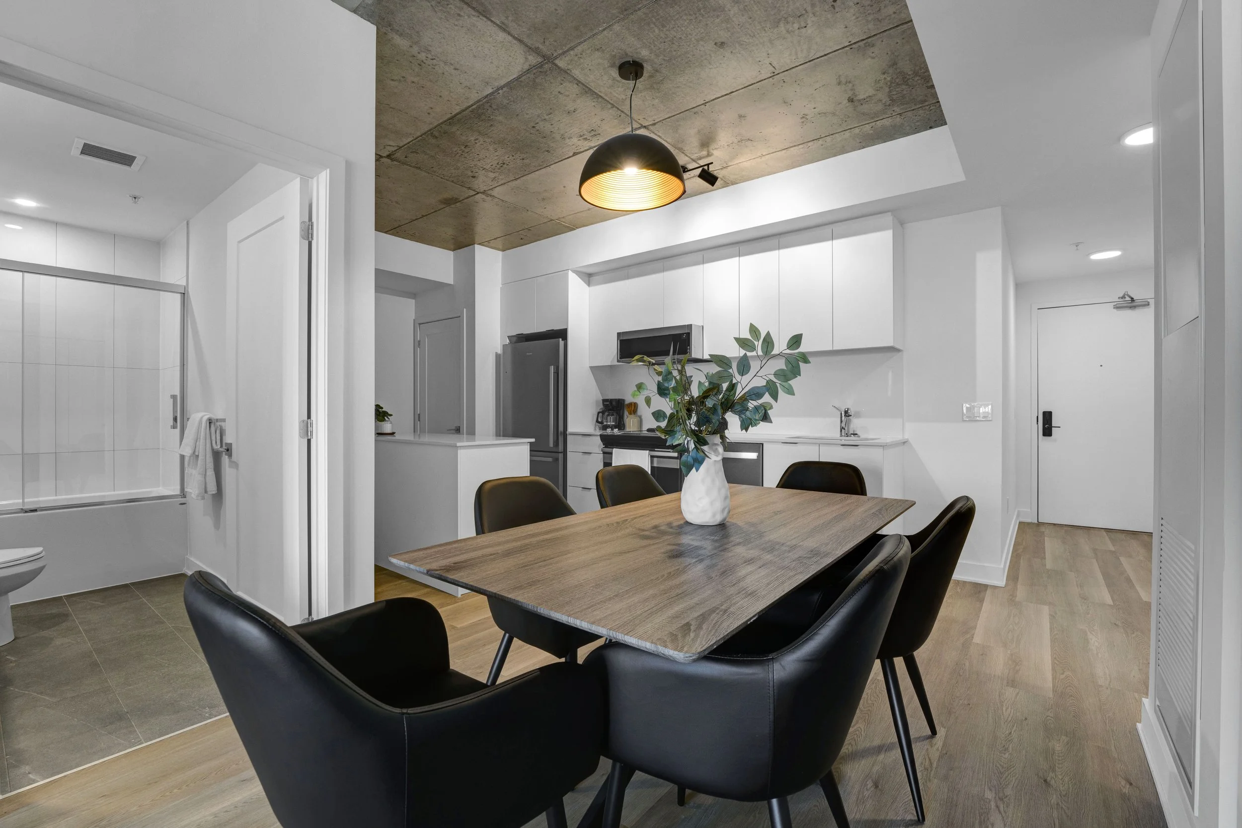 Ottawa Real Estate Photographer Modern kitchen and dining area with a wooden table, black chairs, white cabinets, stainless steel appliances, and a decorative plant in a white vase on the table, with concrete ceiling and bright lighting.