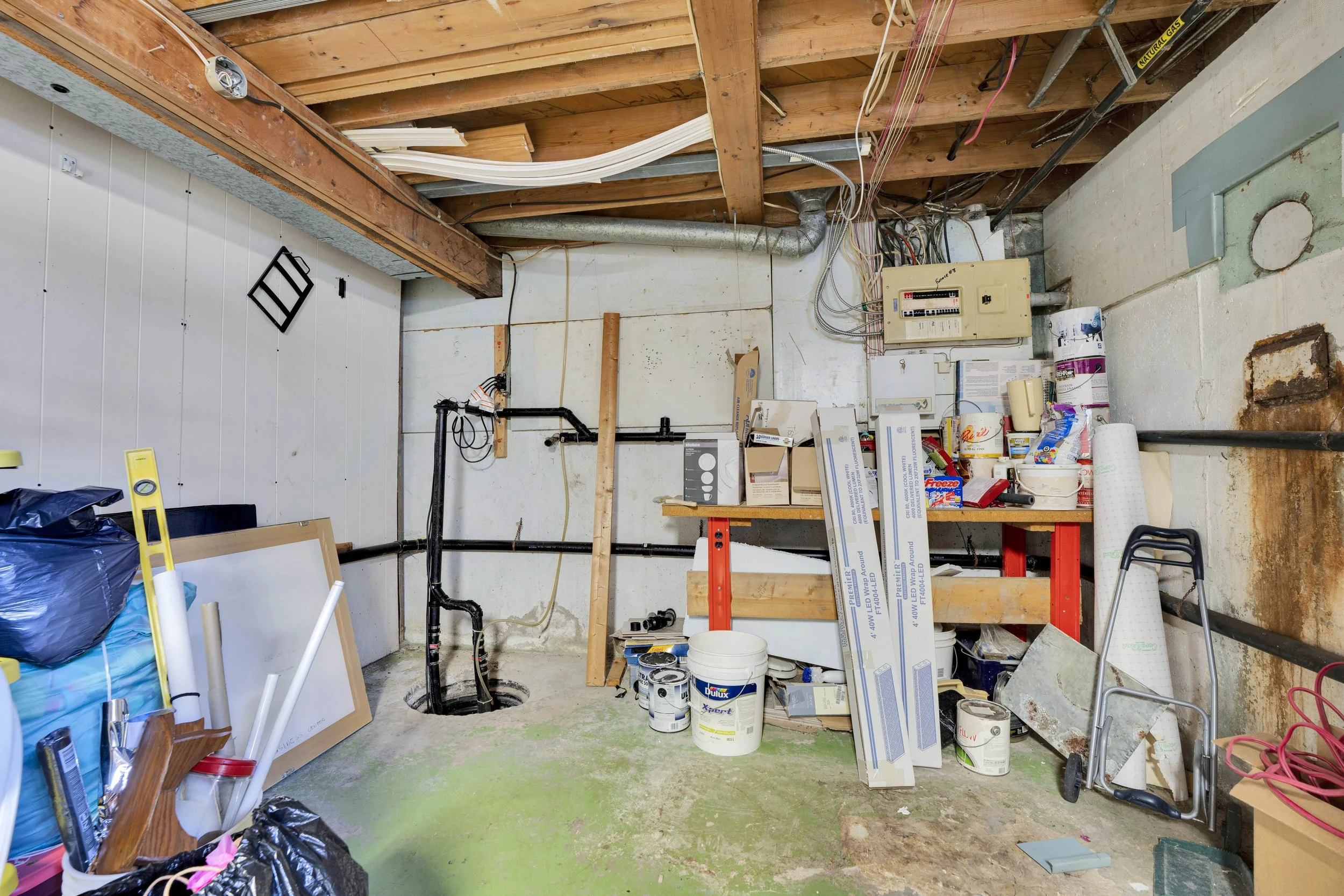 An unfinished basement with exposed wooden beams and electrical wiring. The floor has a greenish tint with construction supplies, paint cans, and boxes scattered around. There's a black pipe, a level, and some boxed lighting fixtures. The wall on the