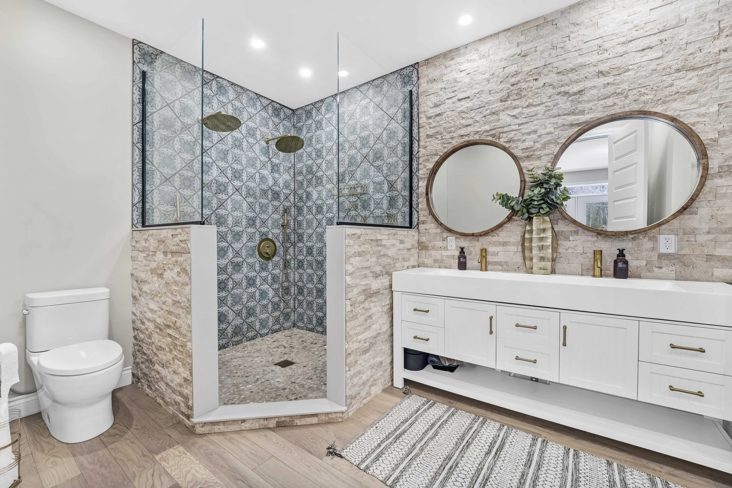 Real Estate Photography in Ottawa Modern bathroom with a walk-in shower, white vanity with gold fixtures, circular mirrors, decorative vase with greenery, wooden flooring, and textured brick wall.