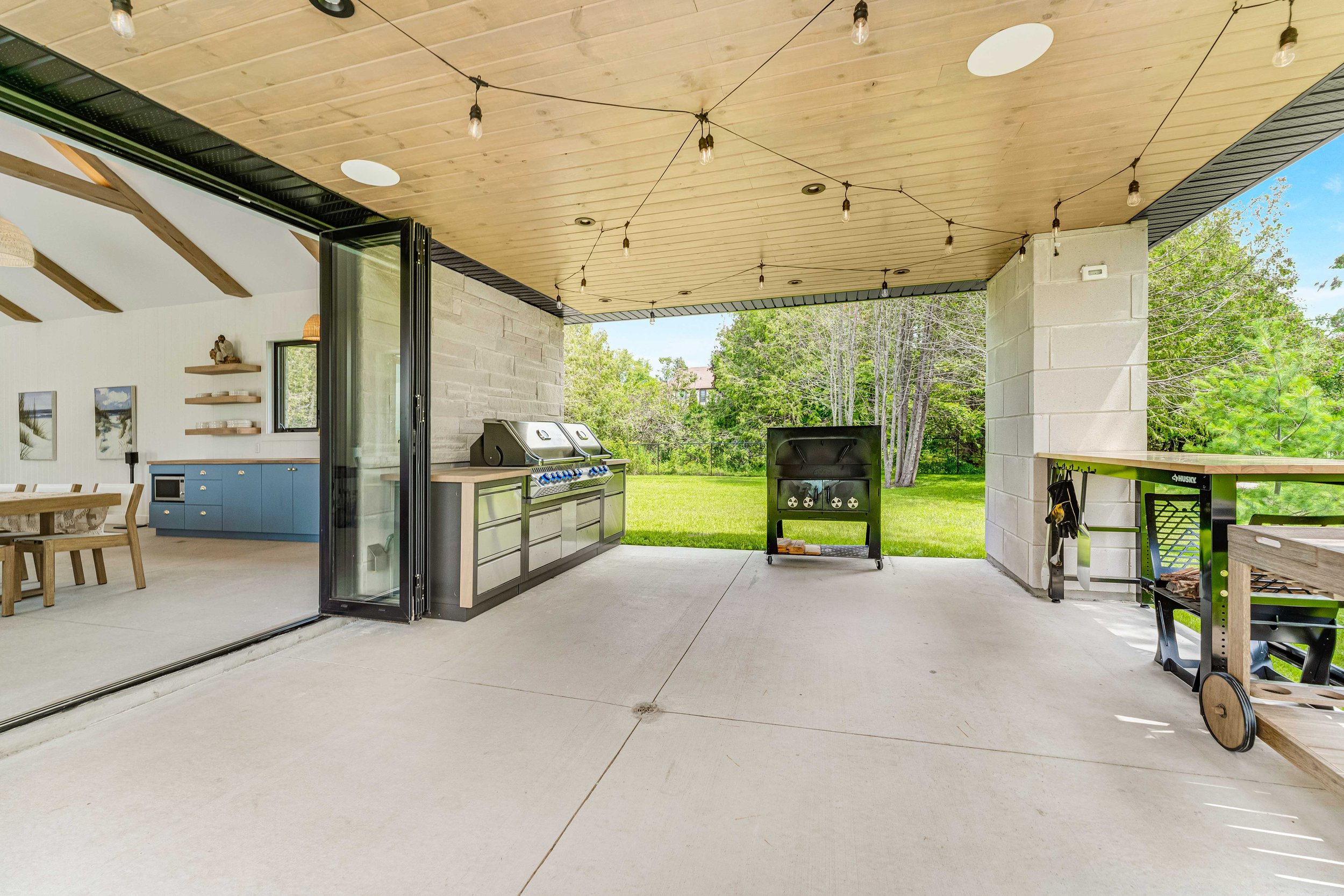 Outdoor covered patio with a BBQ grill, string lights, and a view of a lawn and trees. The patio is connected to an indoor dining area with blue cabinets and a wooden dining table.