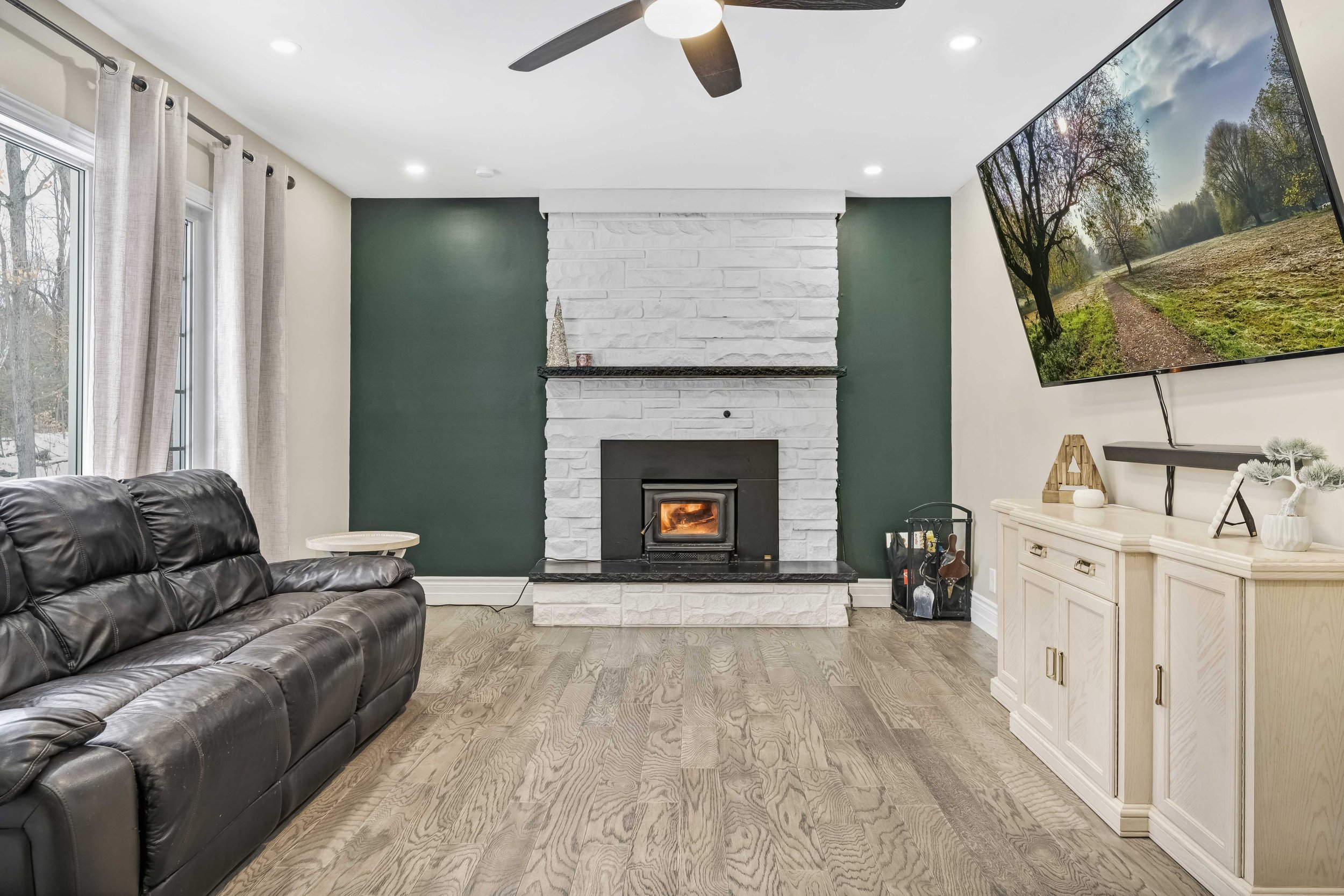 Living room with a white brick fireplace on a green accent wall, a black leather sofa, a side table, a large flat-screen TV mounted on a white wall, and a light-colored cabinet with decorations, hardwood flooring, and curtains on a window. Ottawa