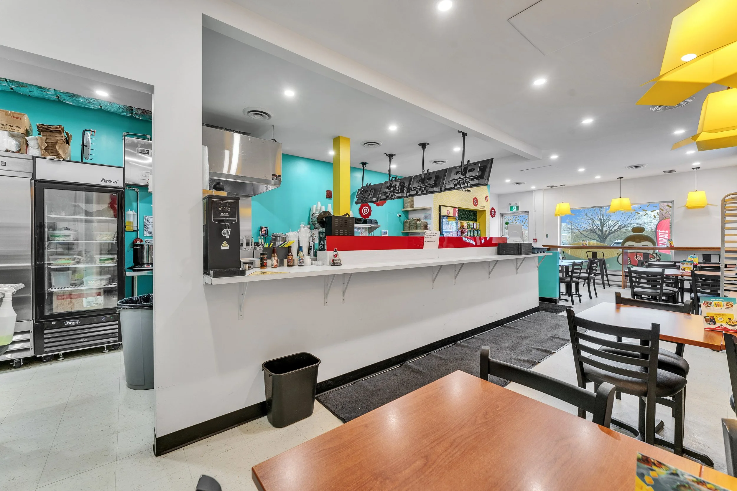 NVISION3D.ca Real Estate Media in Ottawa Interior of a casual restaurant with a brightly colored counter area, black chairs, tables, yellow pendant lights, and large windows showing an outdoor scene.
