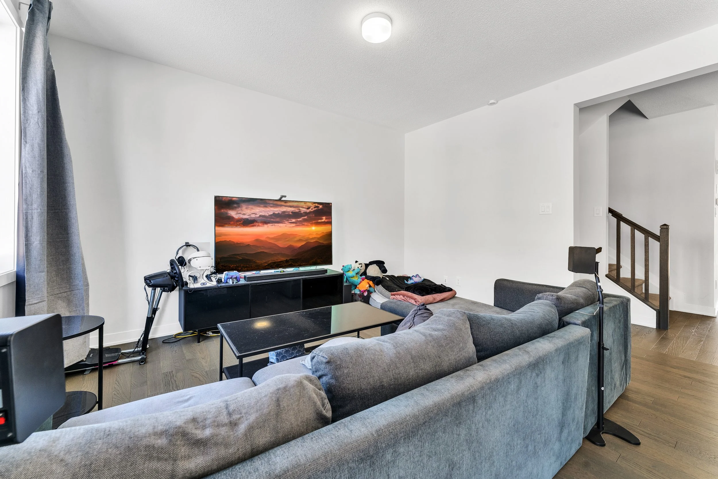 Living room with a gray sectional sofa, a black TV stand with a large flat-screen TV displaying a sunset, and various toys and gadgets on the TV stand. There is a window with gray curtains to the left, and the room has white walls and a wooden floor.