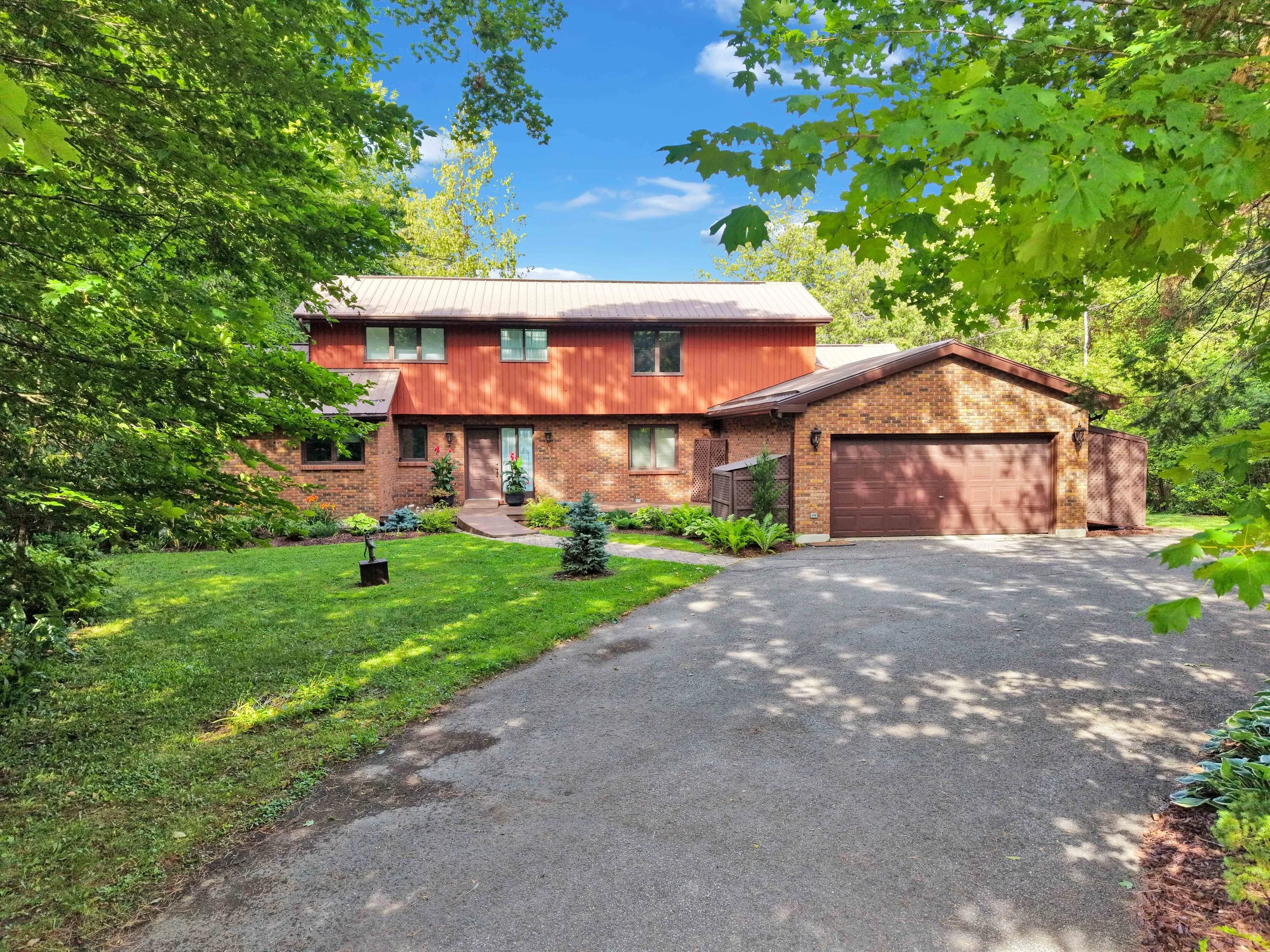 NVISION 3D Real Estate Media  A house with brick and red siding walls, a metal roof, and a brown garage door, surrounded by green trees and plants, with a paved driveway leading to it under a partly cloudy sky.