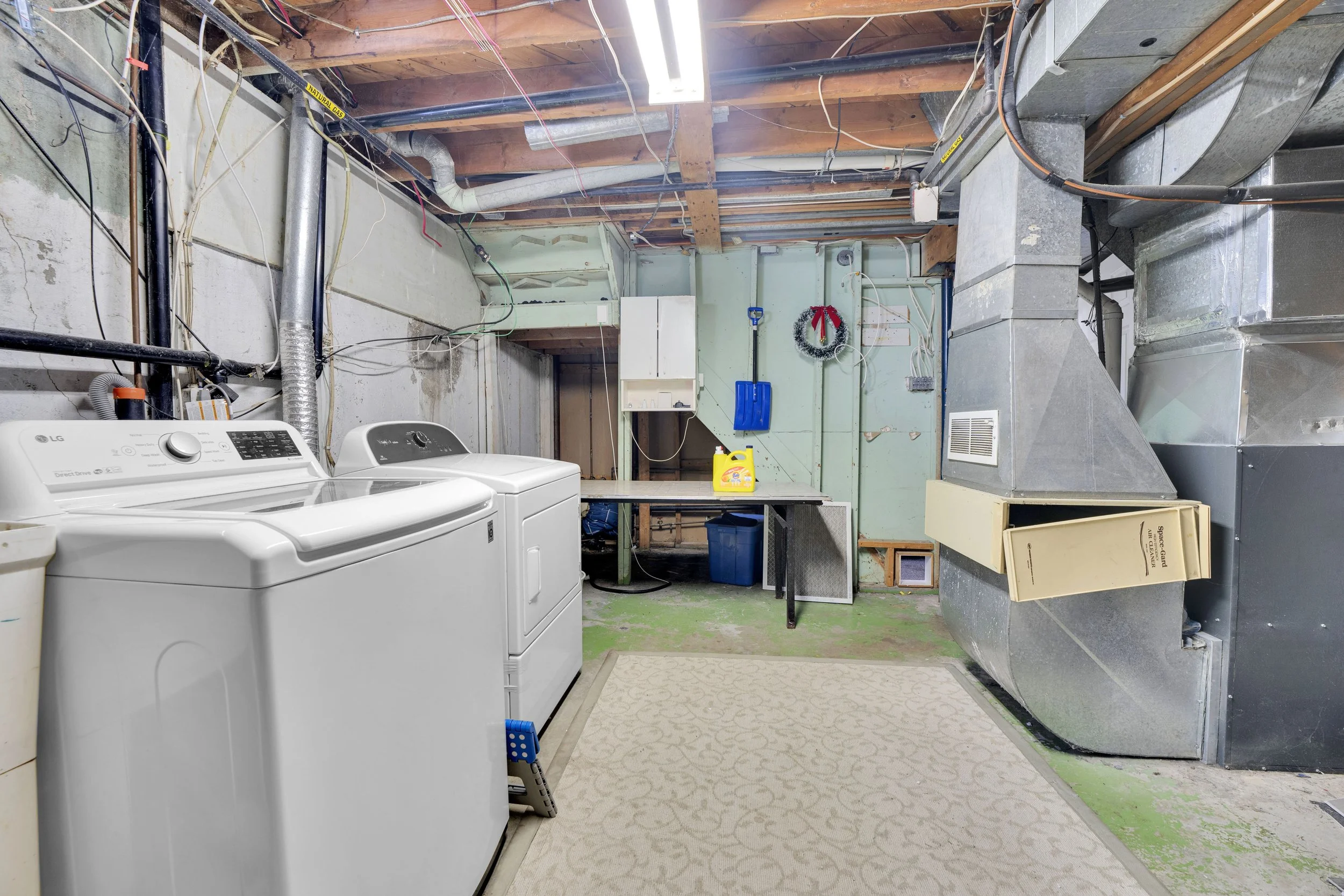 Ottawa Real Estate Media Services  A laundry area with a washing machine and dryer, exposed ceiling with wooden beams and wiring, overhead fluorescent light, and a utility vent. There are shelves, a blue bin, and a beige box on the floor.